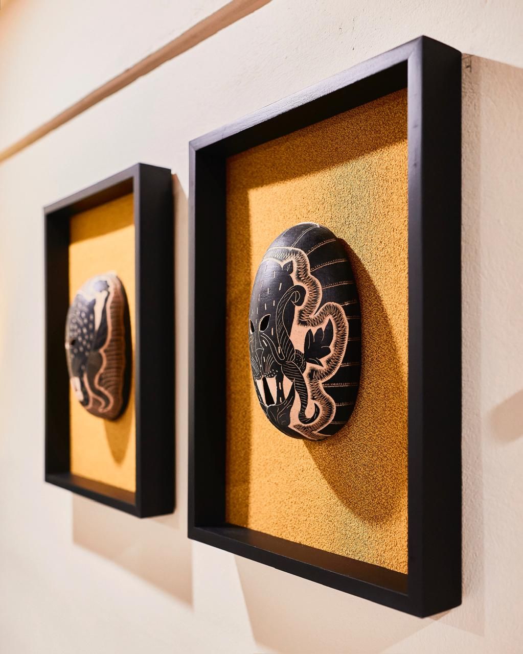 Two framed, oval-shaped art pieces with black frames and yellow backgrounds, mounted on a cream-colored wall.