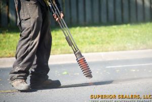 Worker using a plate compactor to smooth asphalt. He wears a safety vest, working on a road near a curb.