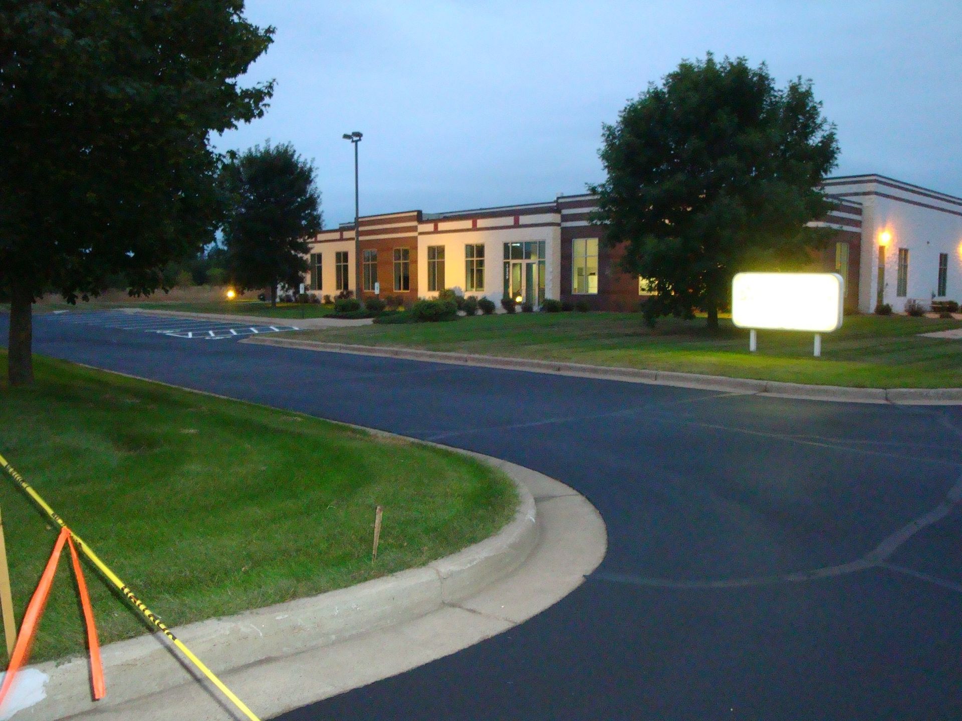 Building with sign, trees, and curved road. Dark asphalt and green grass under a dusk sky.