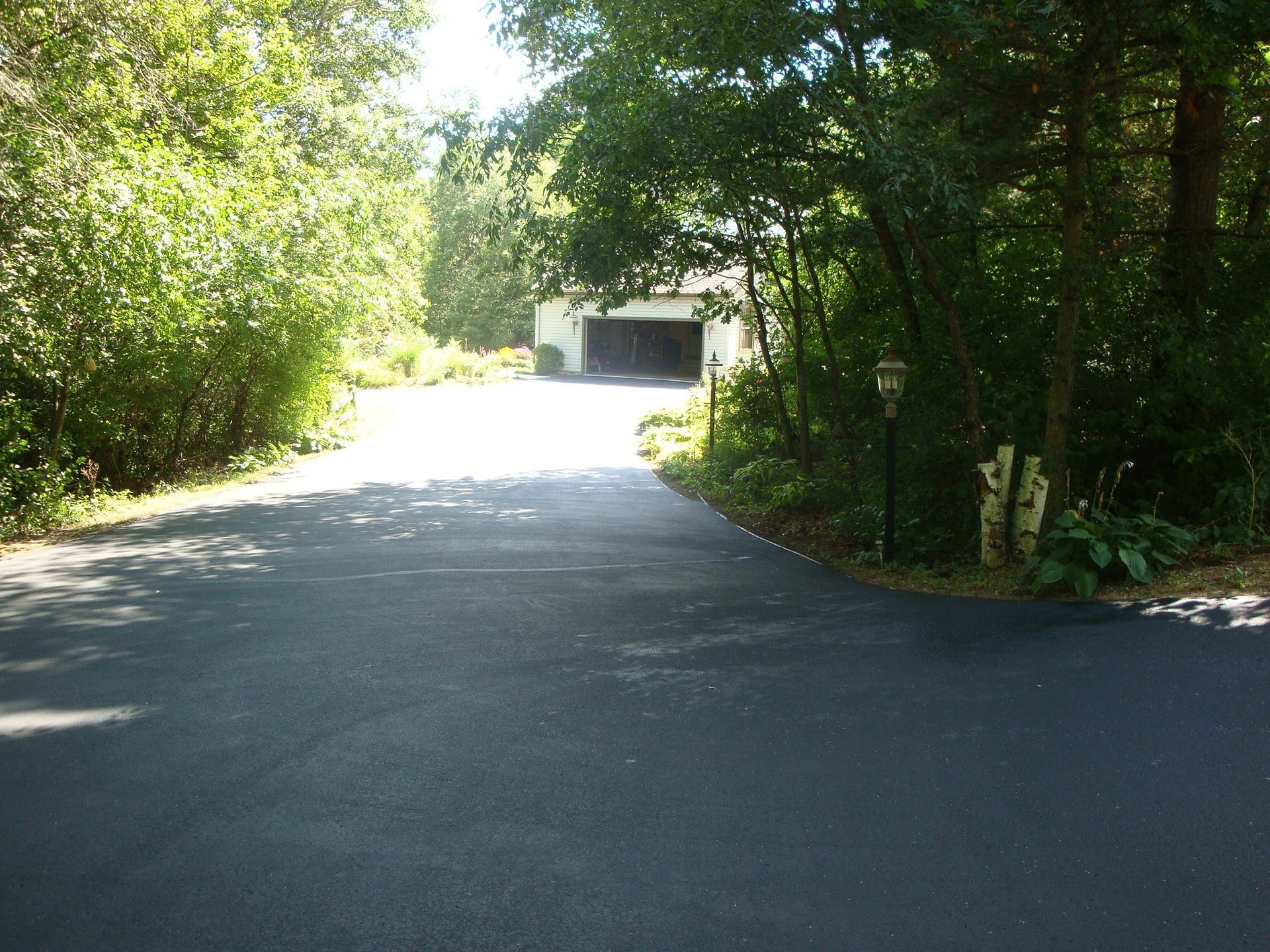 Asphalt driveway leading to a two-car garage, flanked by trees and greenery. Bright sunlight.