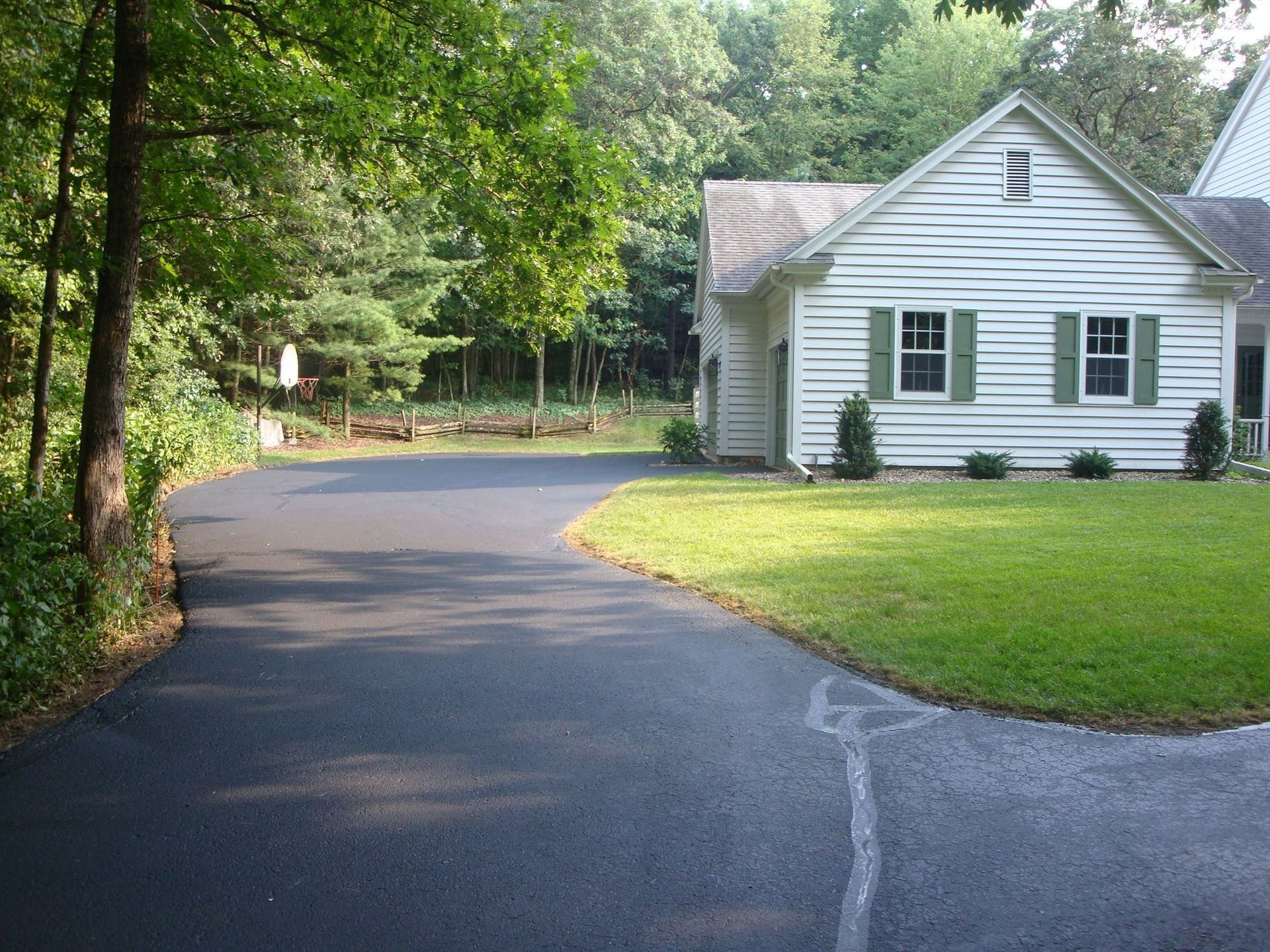 Asphalt driveway curving to a white house with green shutters, surrounded by trees and grass.