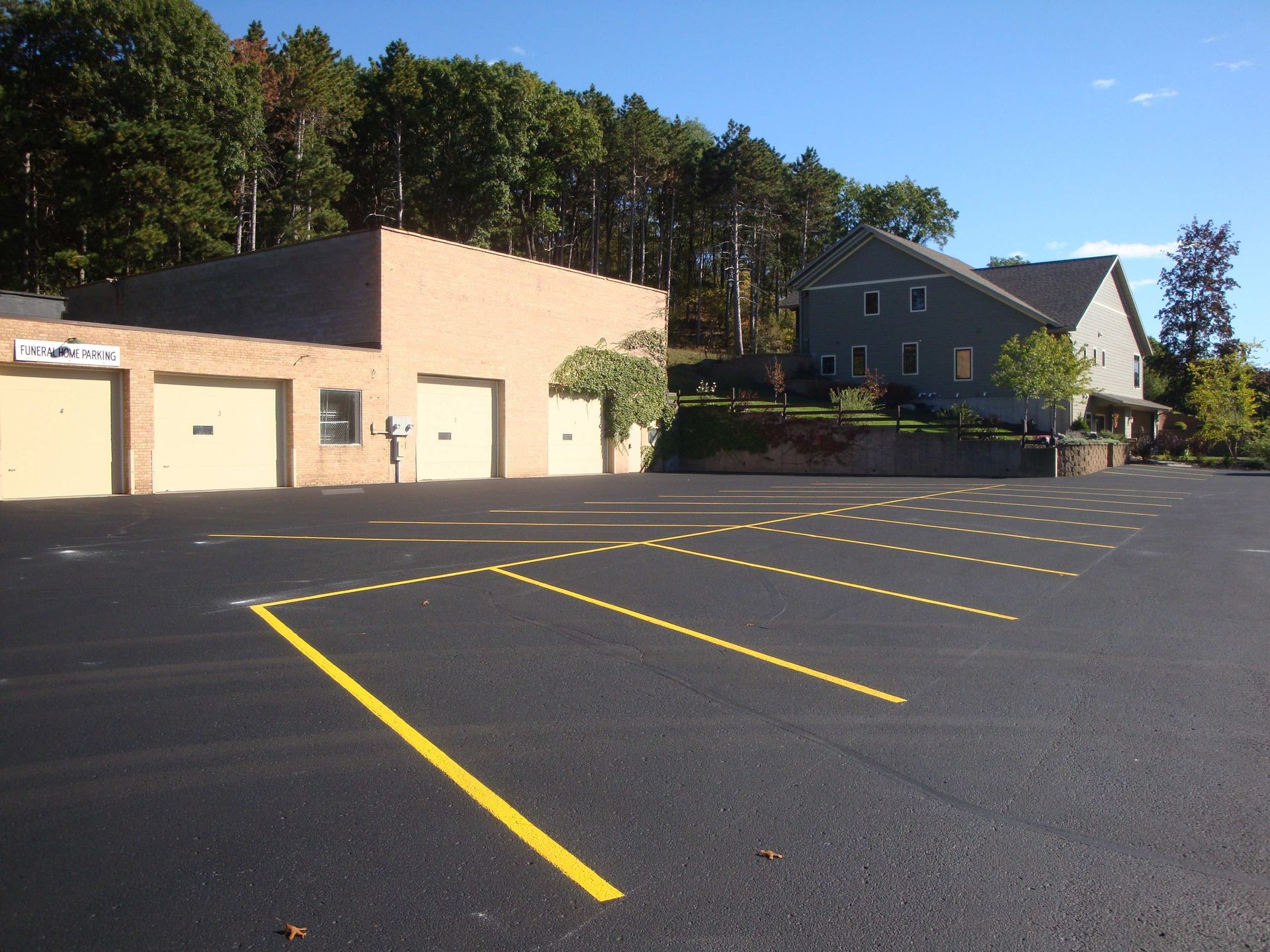 Newly paved parking lot with yellow lines in front of a beige building and a two-story house.