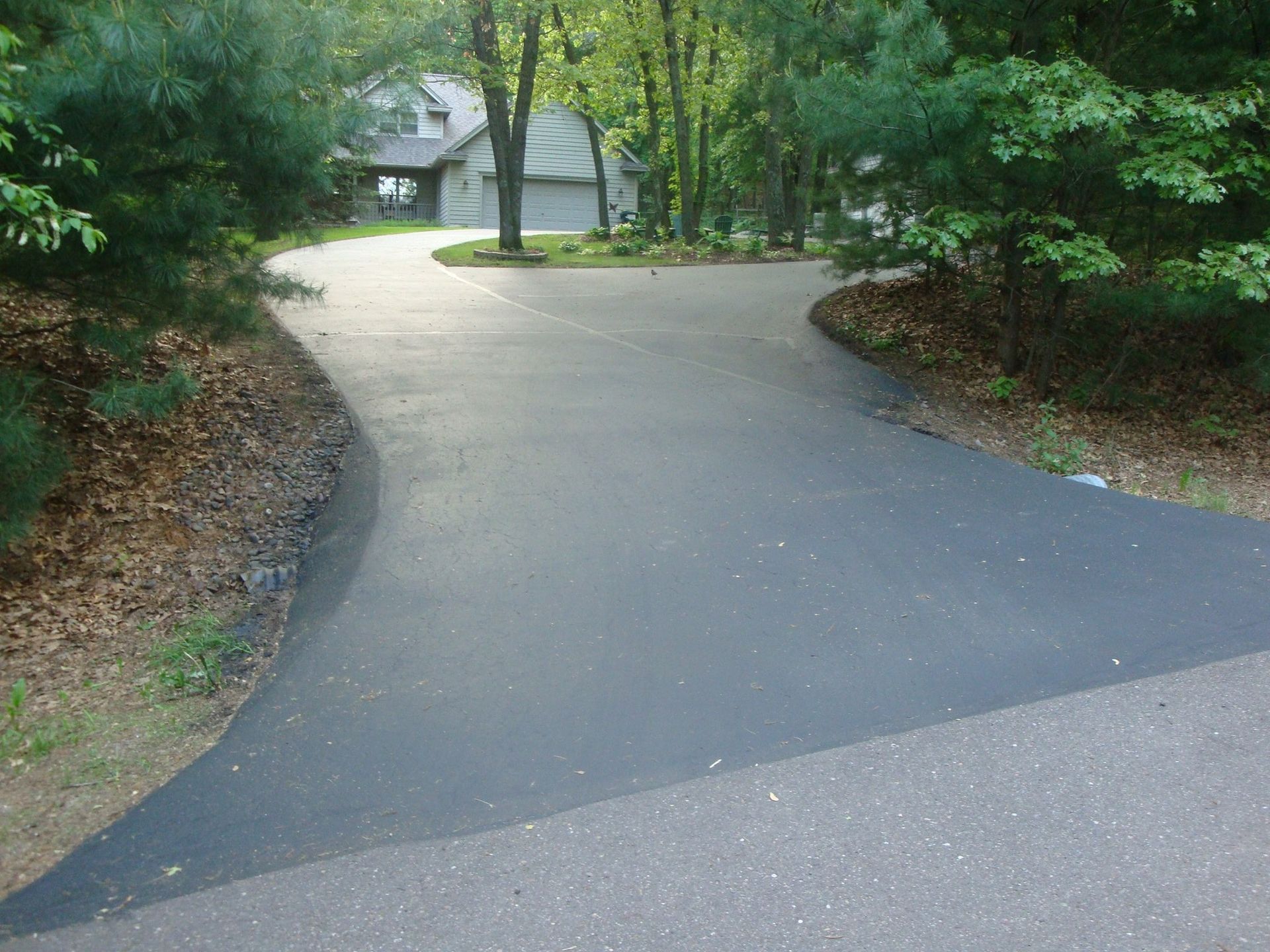 Black asphalt driveway curves toward a house in a wooded area.