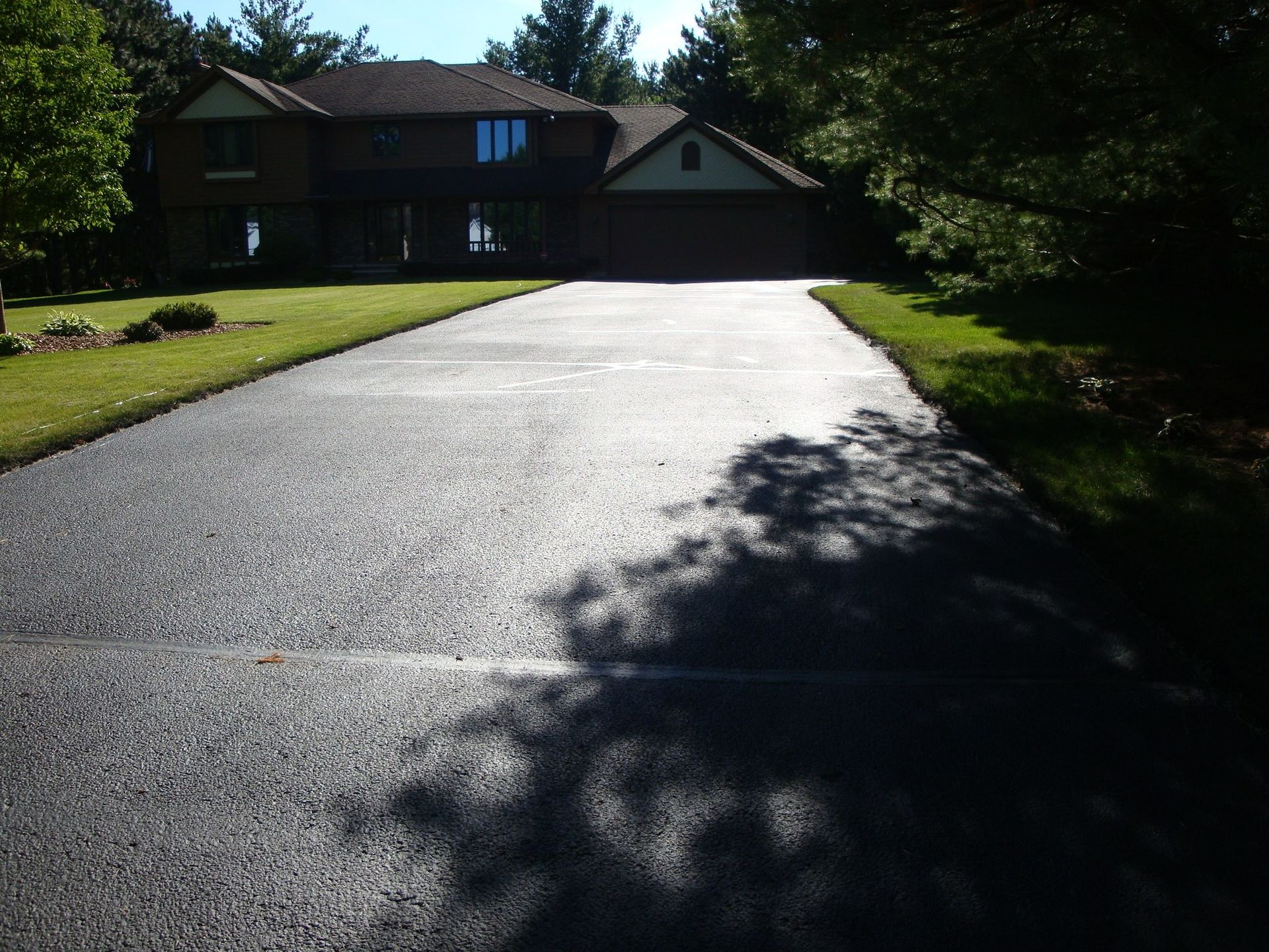 Asphalt driveway leading to a two-story brick house with a green lawn on either side. The driveway is shaded.