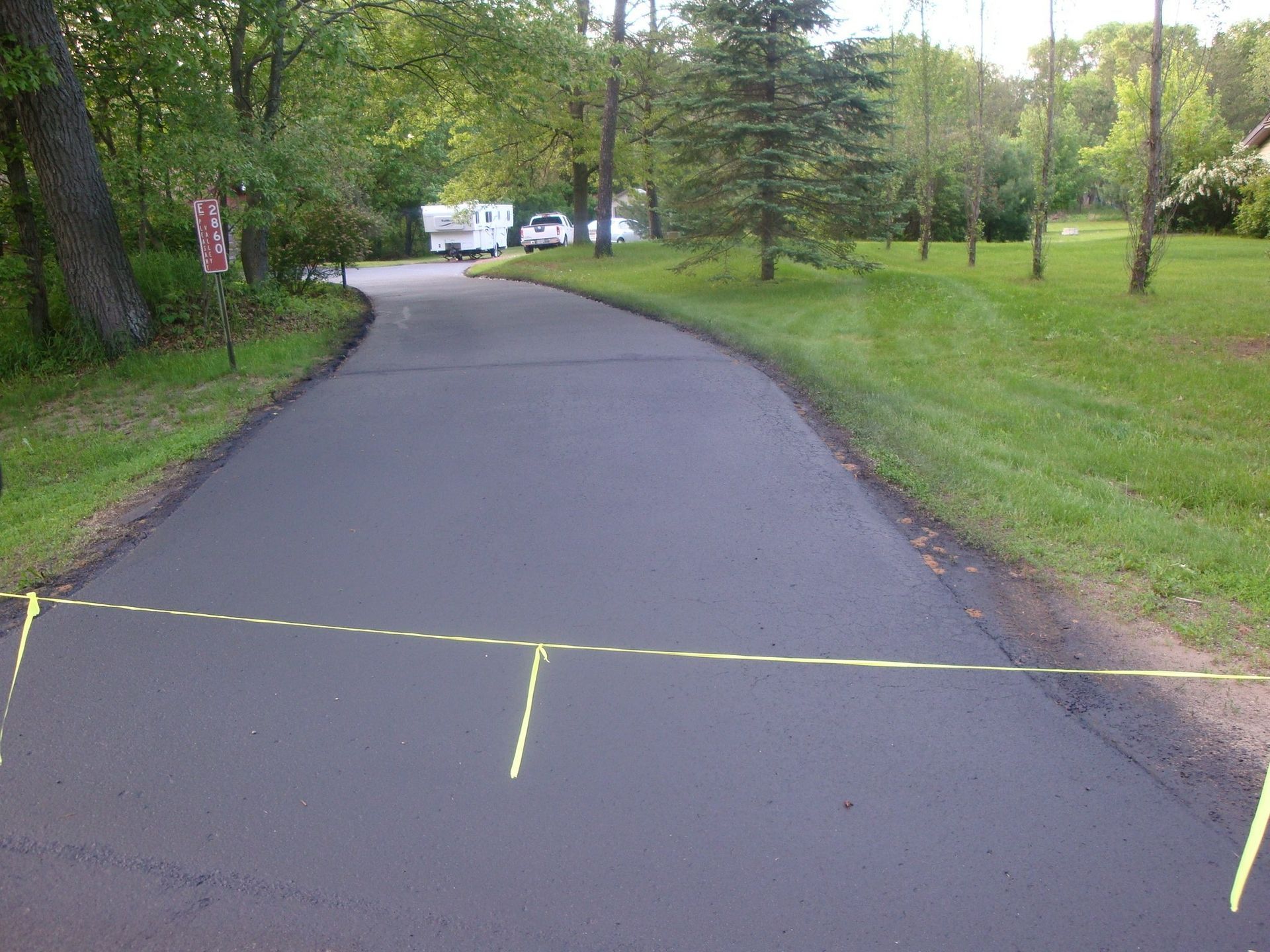 Newly paved asphalt driveway with yellow caution tape, trees, and a trailer.