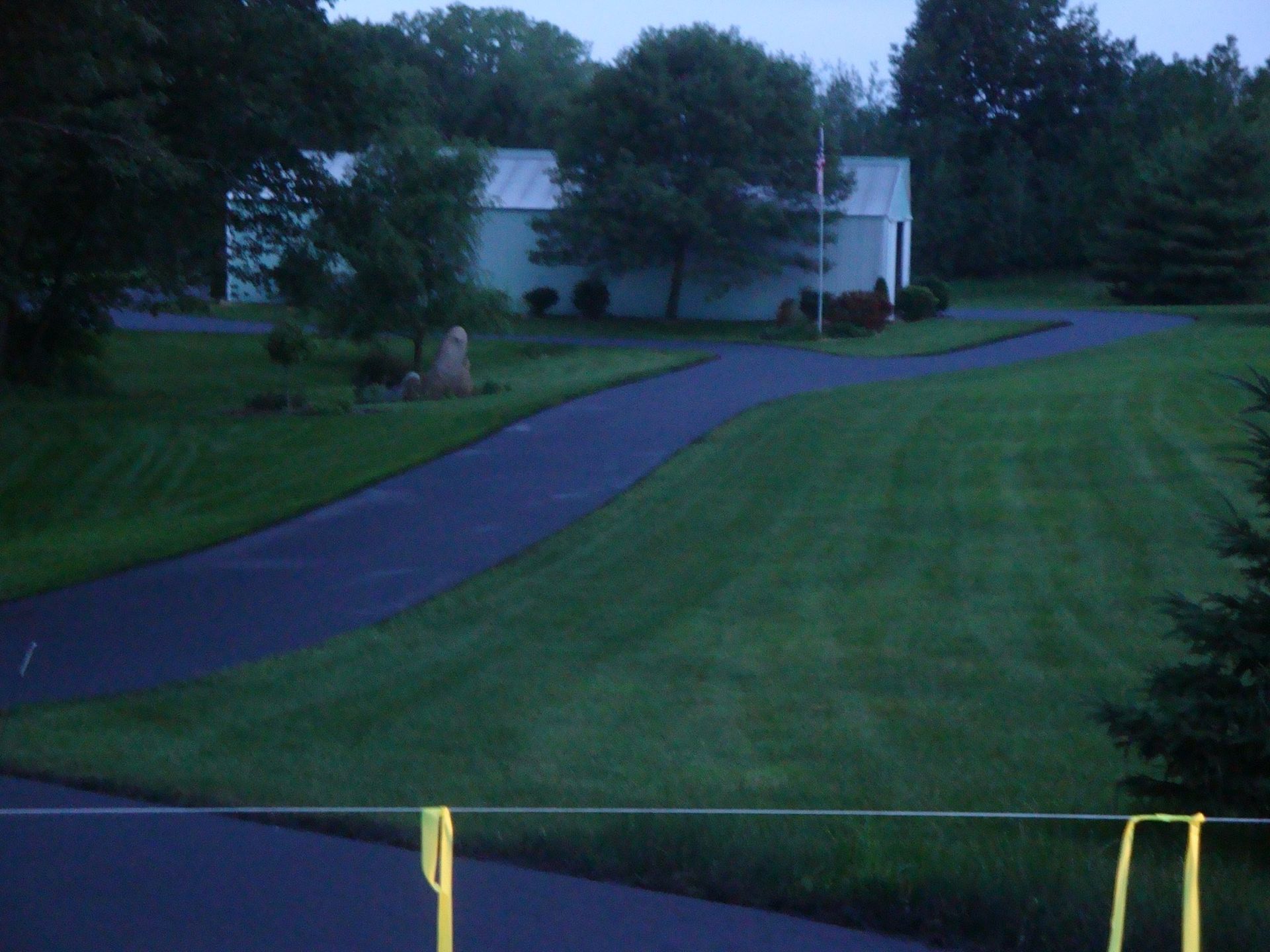 Asphalt driveway curves toward a light blue building with trees in a grassy yard.