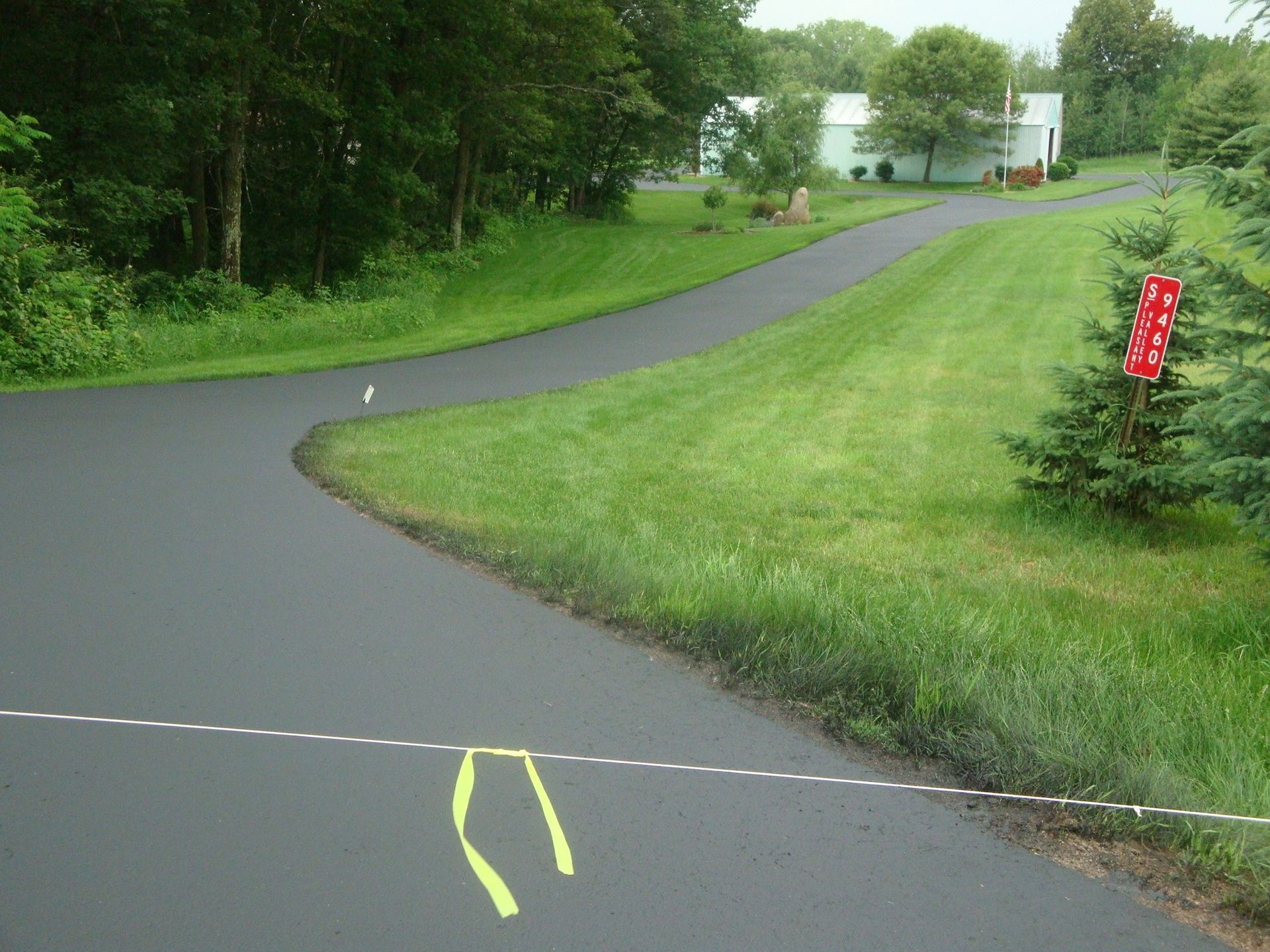 Asphalt driveway curving through grassy yard towards a house in the distance; a red banner hangs from a tree.