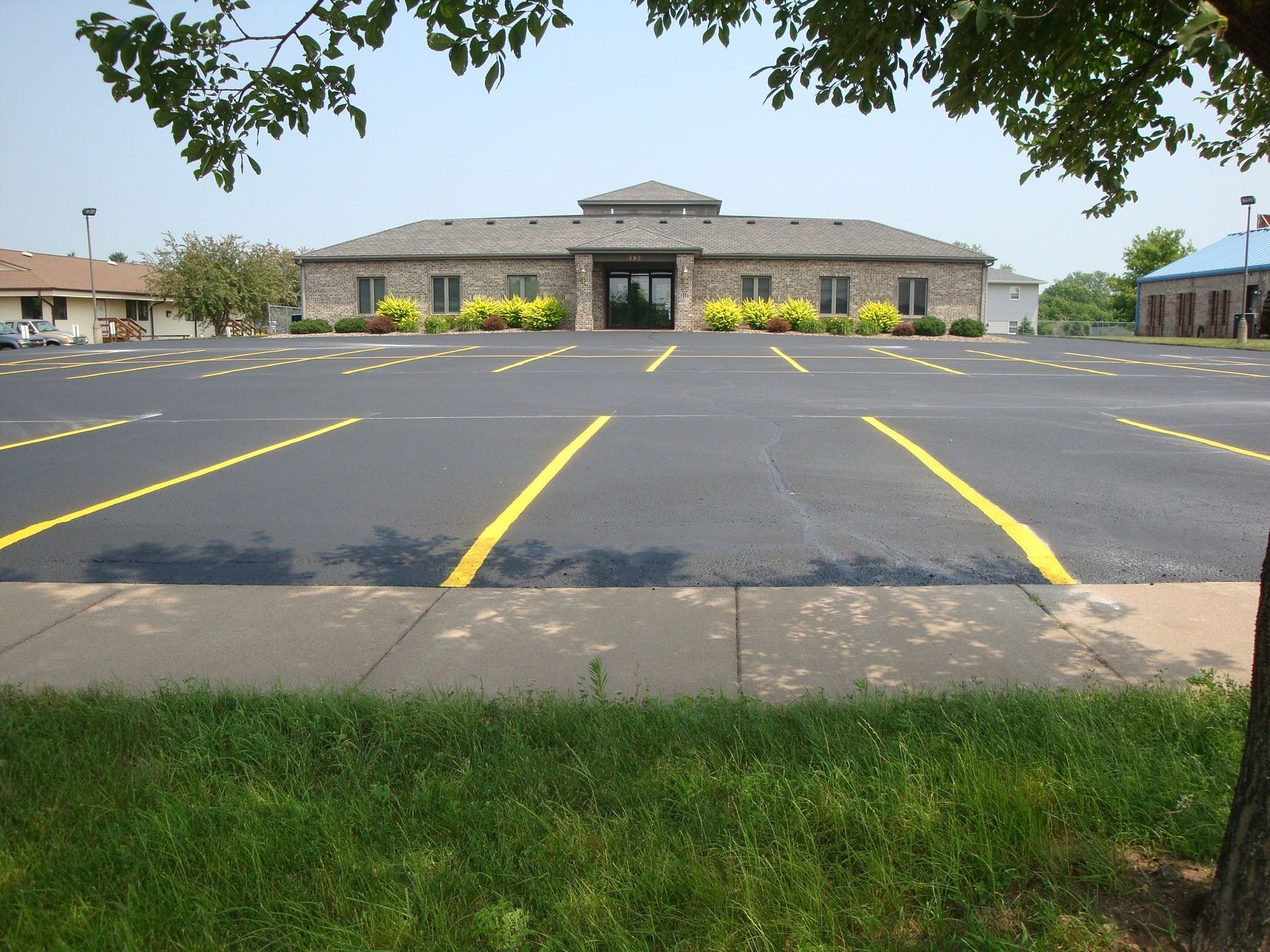 Empty asphalt parking lot in front of a one-story stone building. Yellow parking lines; green grass in foreground.