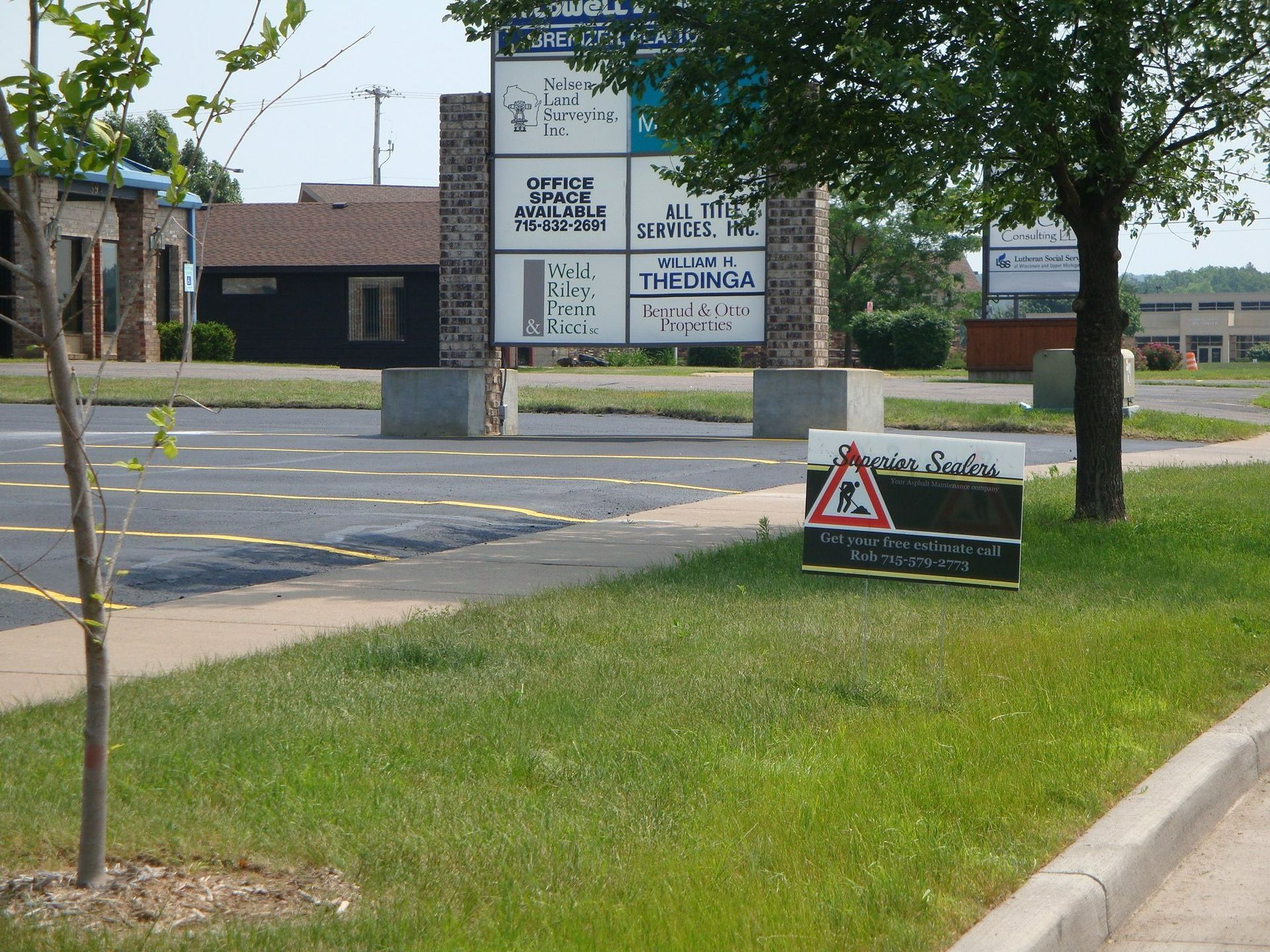 Sign in a grassy area with building in background; parking lot, brown sign, and tree.
