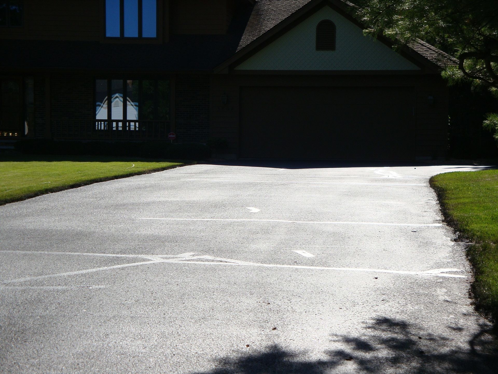 A dark asphalt driveway in front of a two-story house with a garage. Cracks are visible on the driveway's surface.