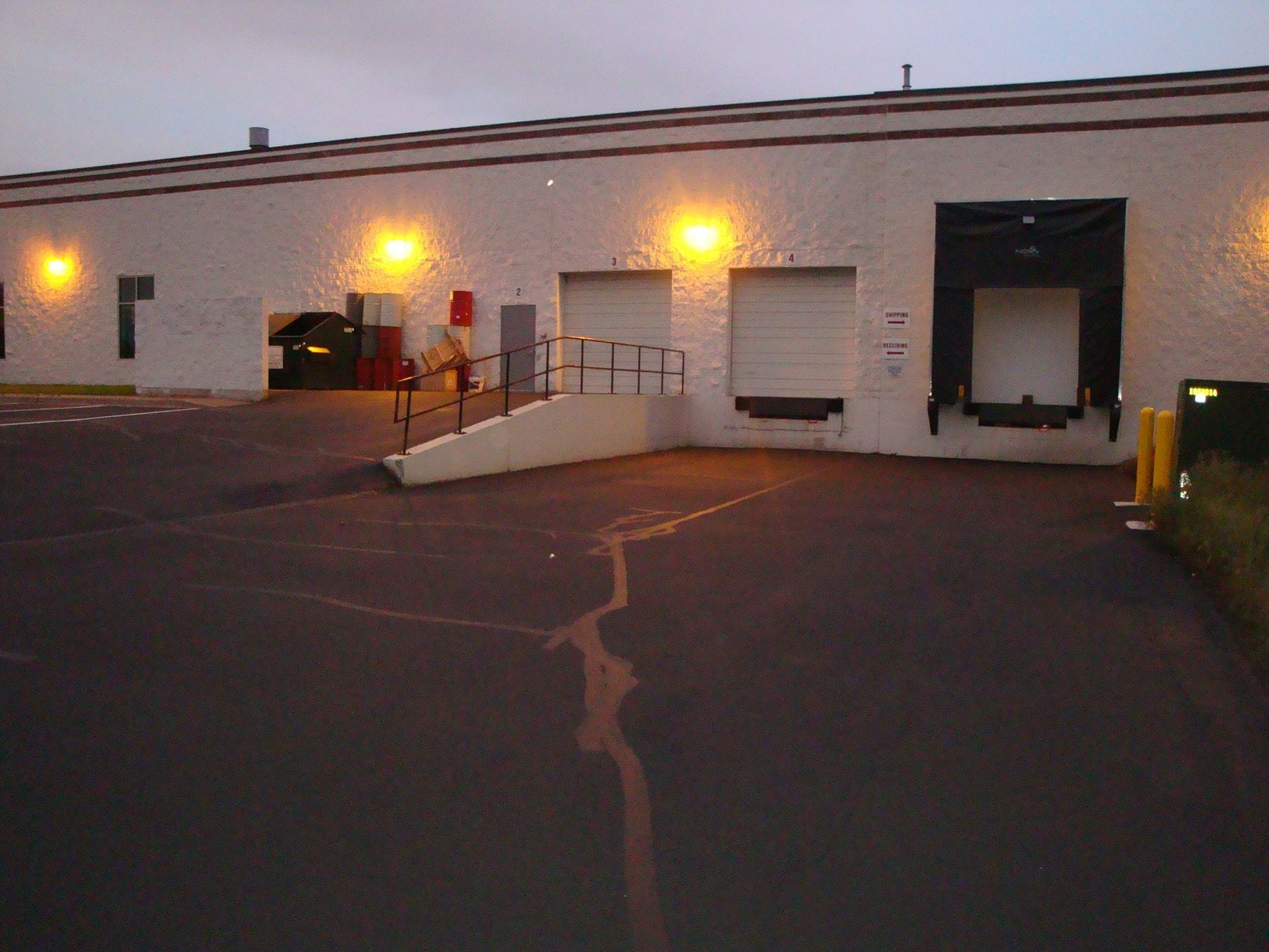 Warehouse exterior with loading docks, lit by orange lights. Asphalt pavement in foreground.