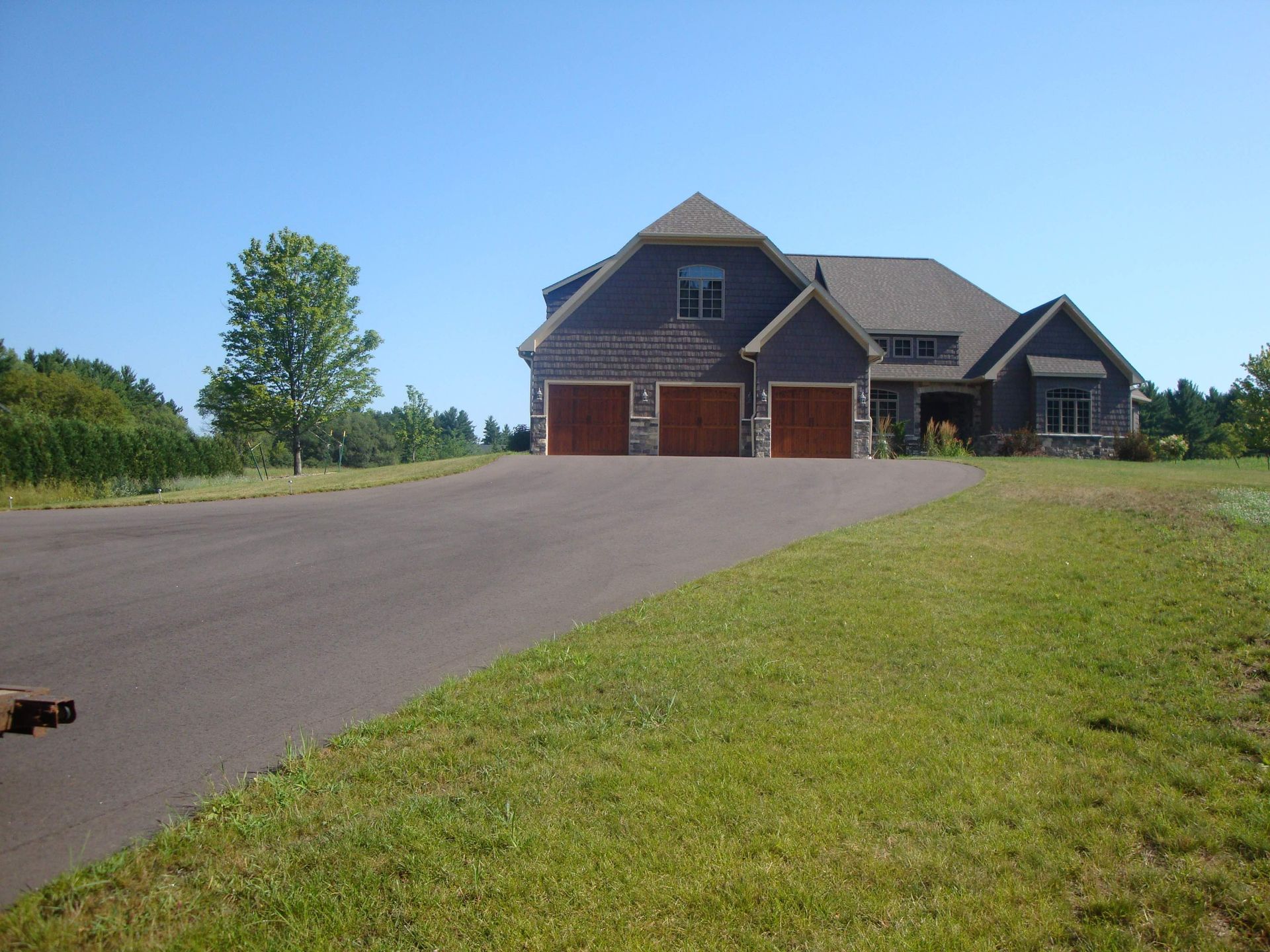 House with asphalt driveway, three-car garage, brown doors, green lawn, and blue sky.