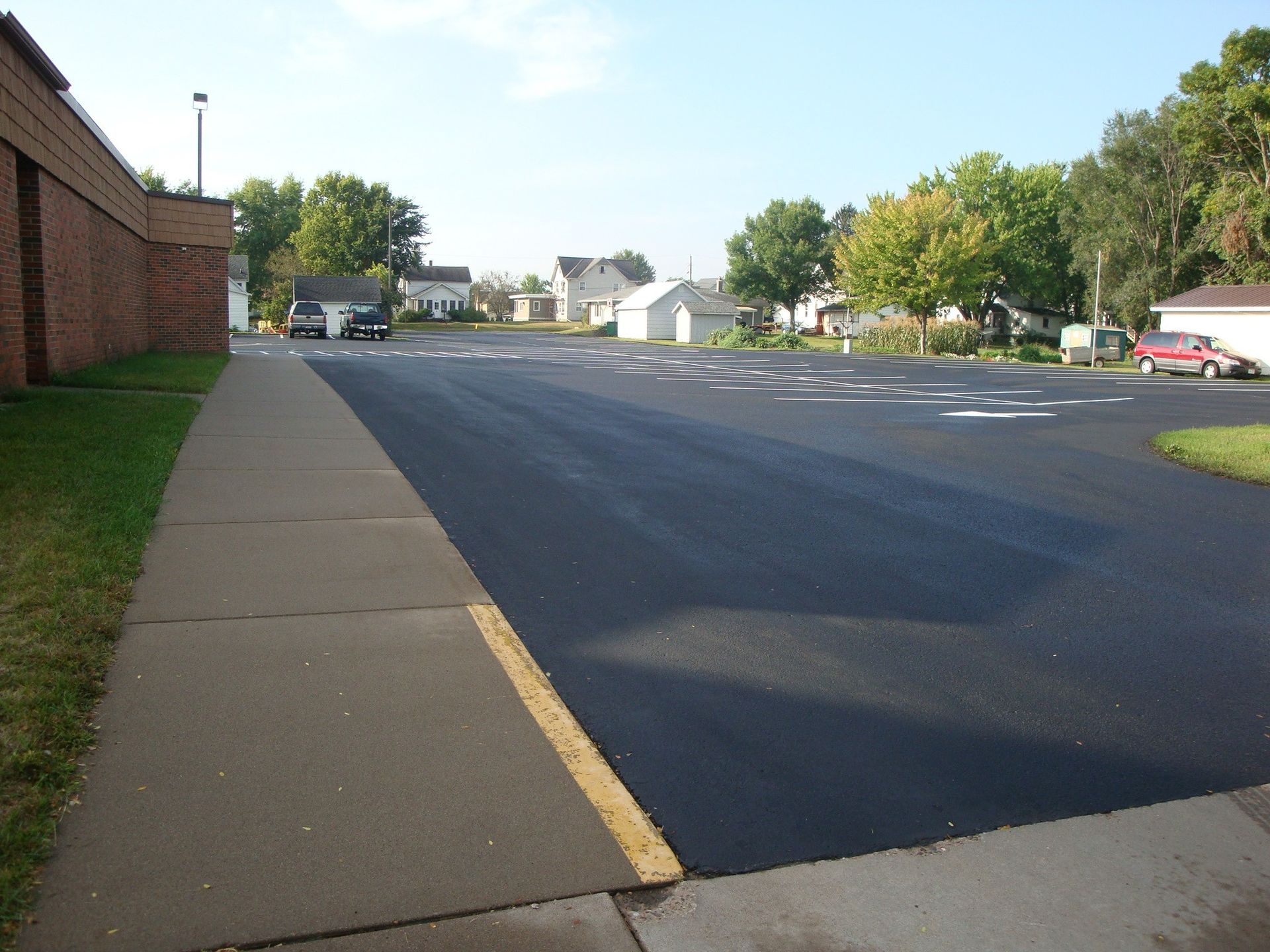 Asphalt parking lot next to a brick building, with a sidewalk on the left, and houses in the background.