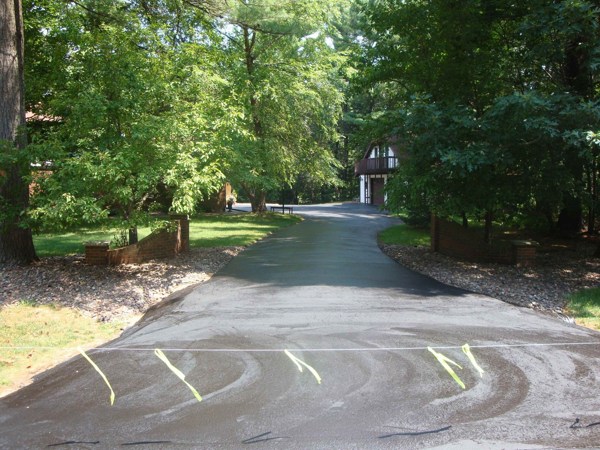 Asphalt driveway curving toward a house, marked with yellow paint. Trees and lawn flank the road.