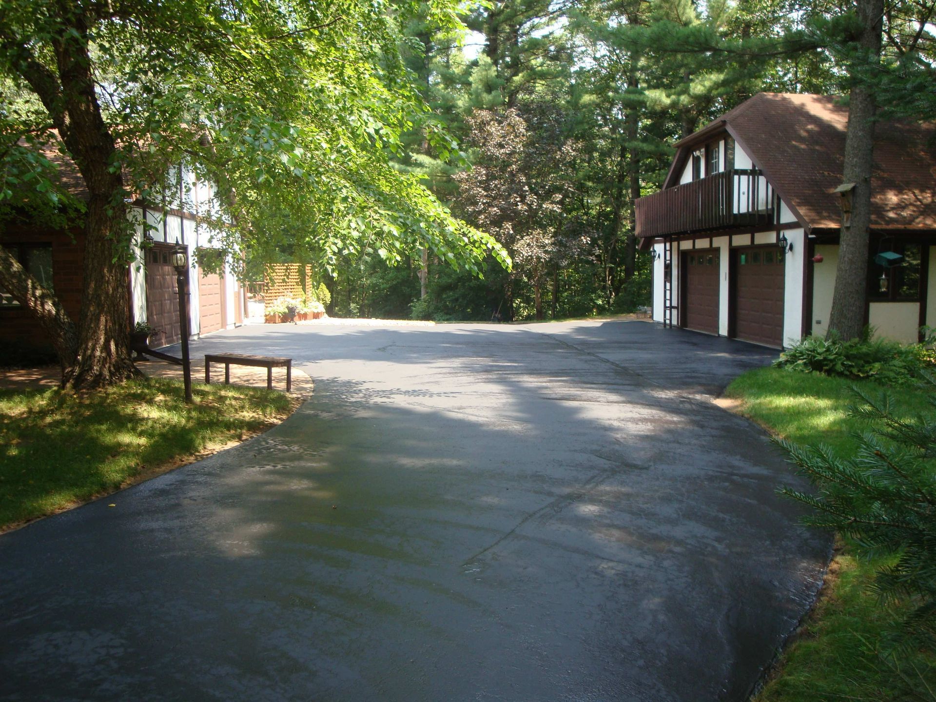 Paved driveway leading to a house with a garage, surrounded by trees. Sunny day.