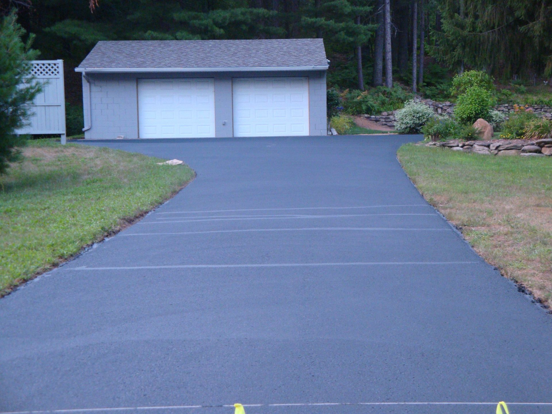 Newly paved, dark gray asphalt driveway leading to a two-car garage. Green grass flanks the driveway.