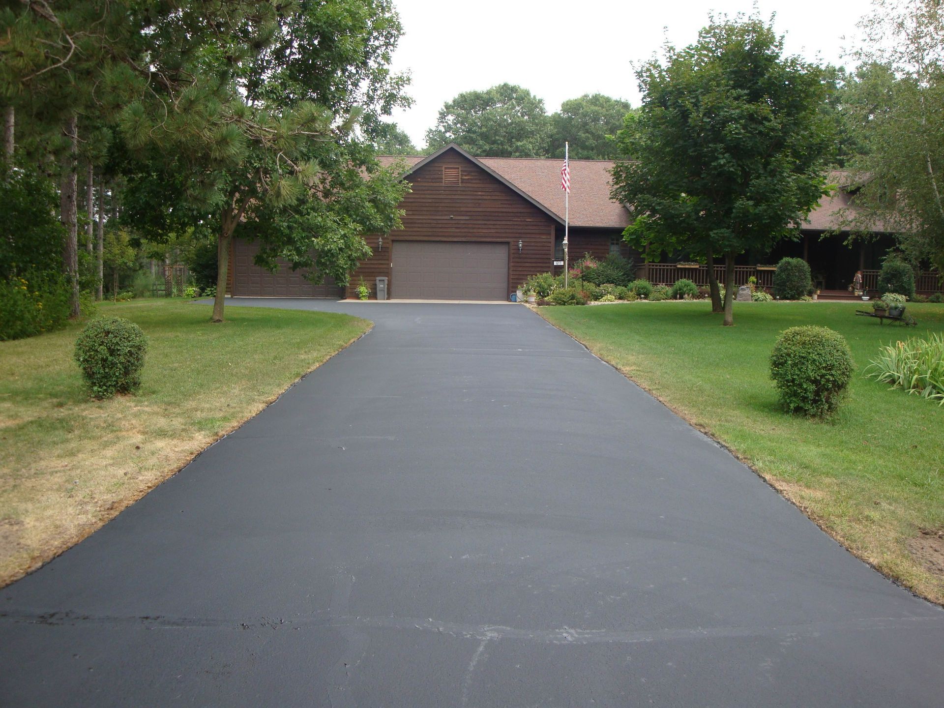 Black asphalt driveway leading to a brown house with a garage, set in a green lawn.