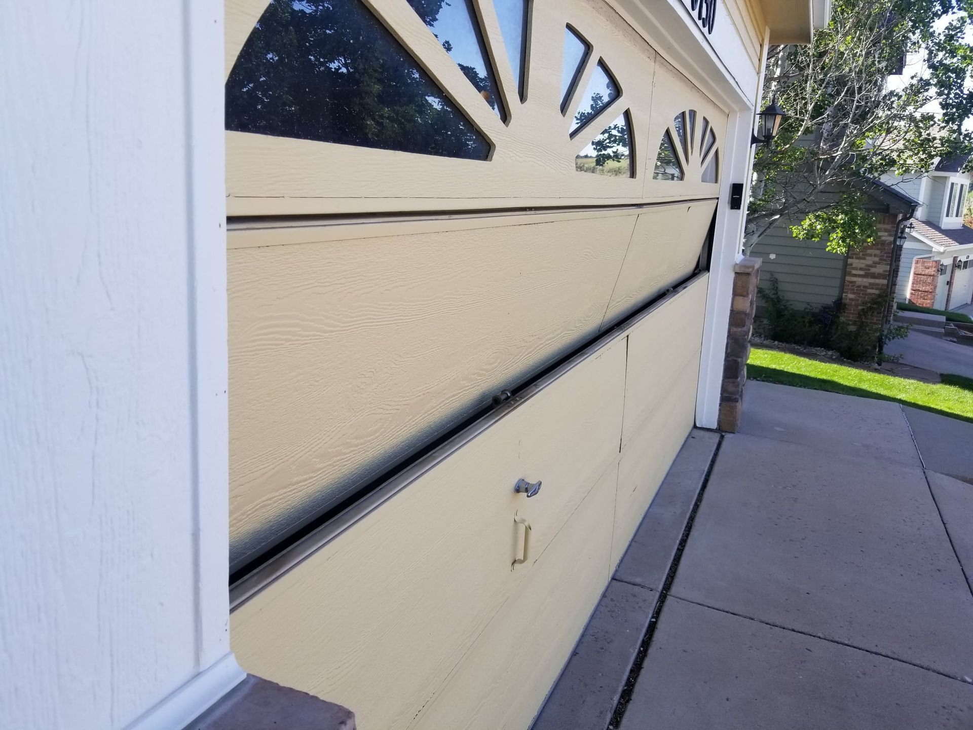 Beige garage door partially open, revealing a crack. A sidewalk and house are in the background.