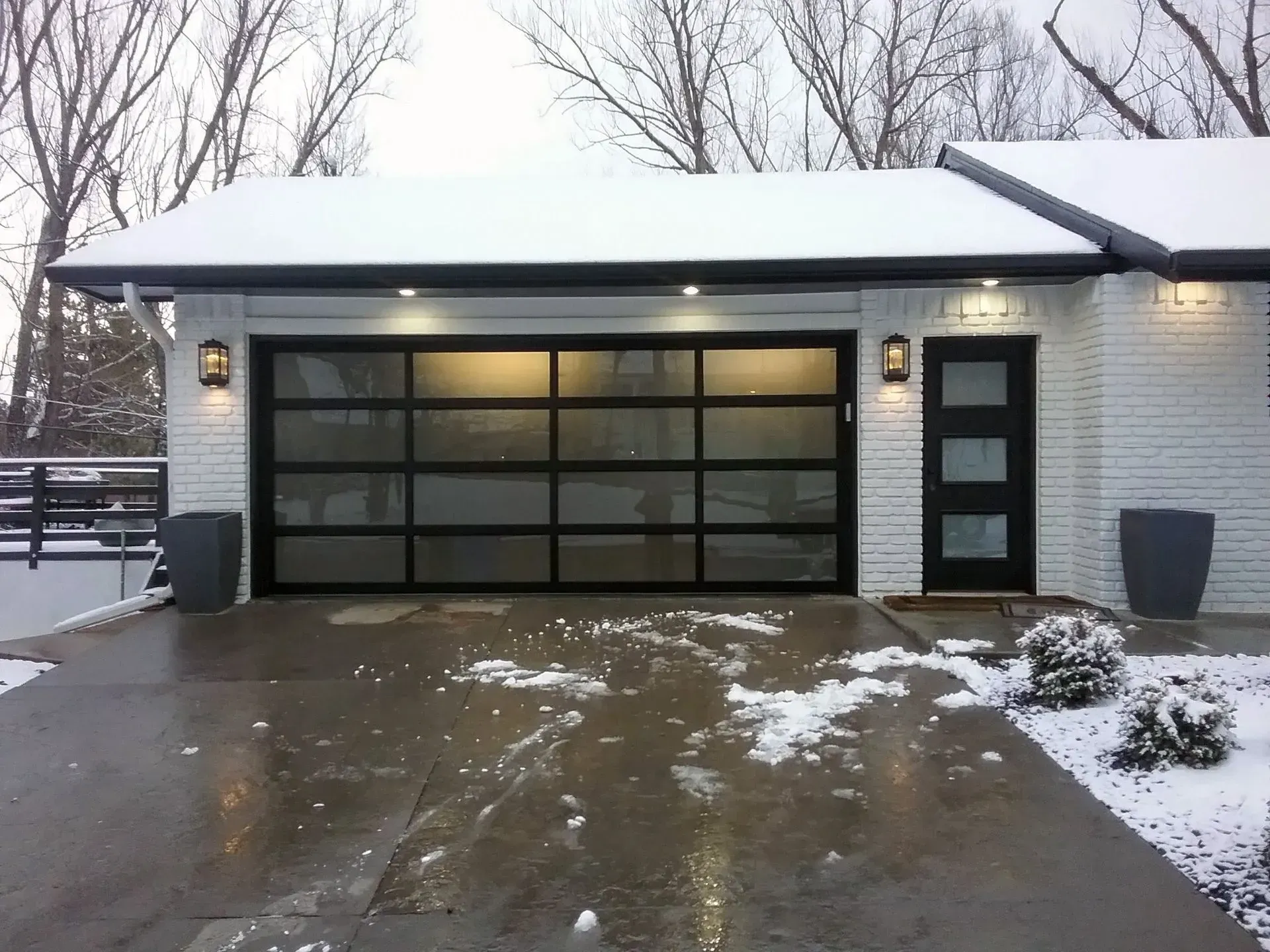 Modern home exterior with snow-covered driveway, glass garage door, black front door, and white brick.