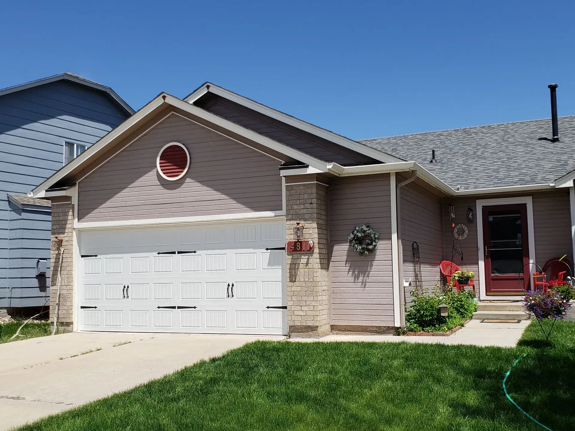House exterior with white garage door, brown siding, red door, and green lawn under a blue sky.