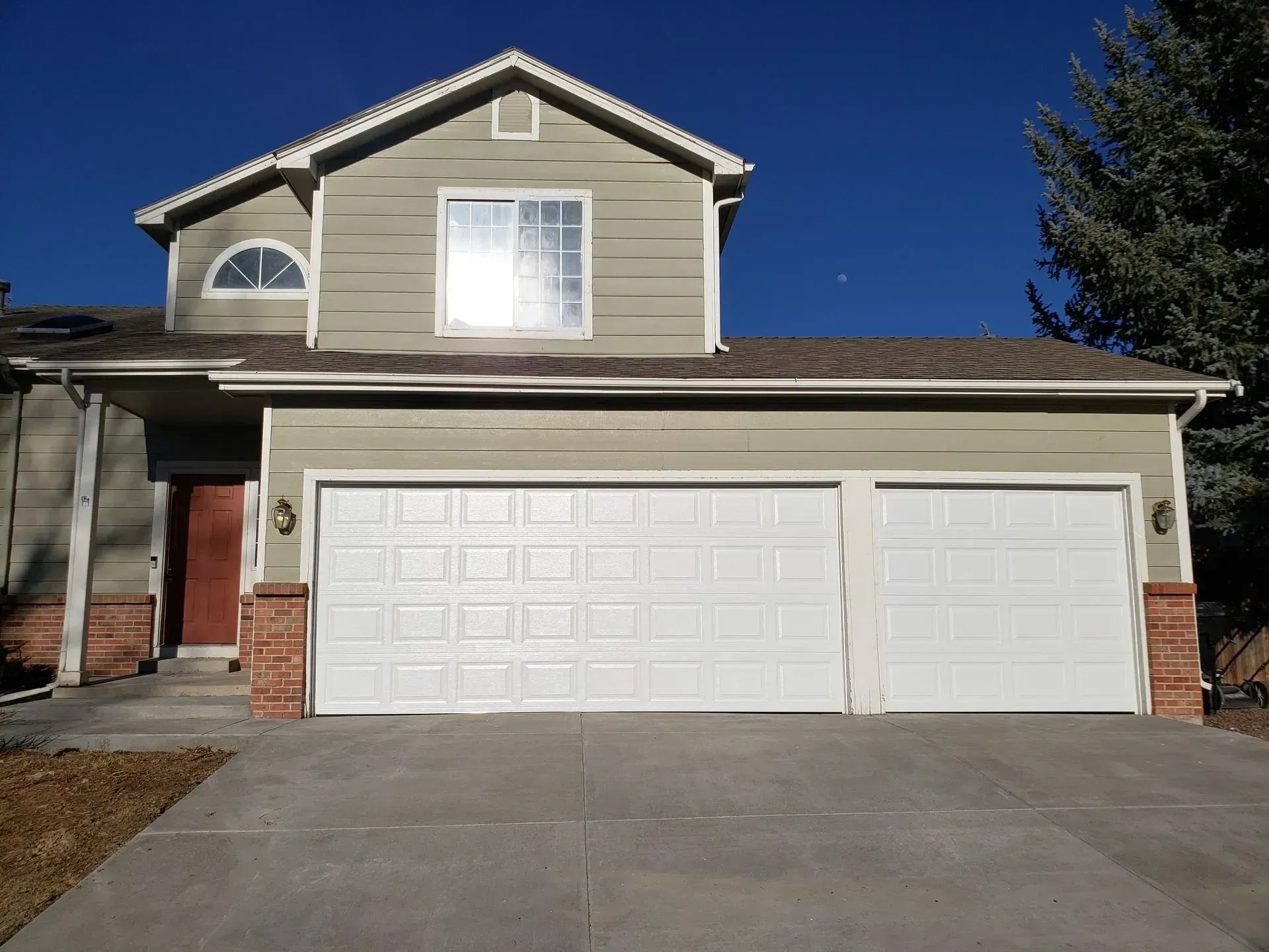Two-story house with a white garage door, olive green siding, red brick accents, and a blue sky.