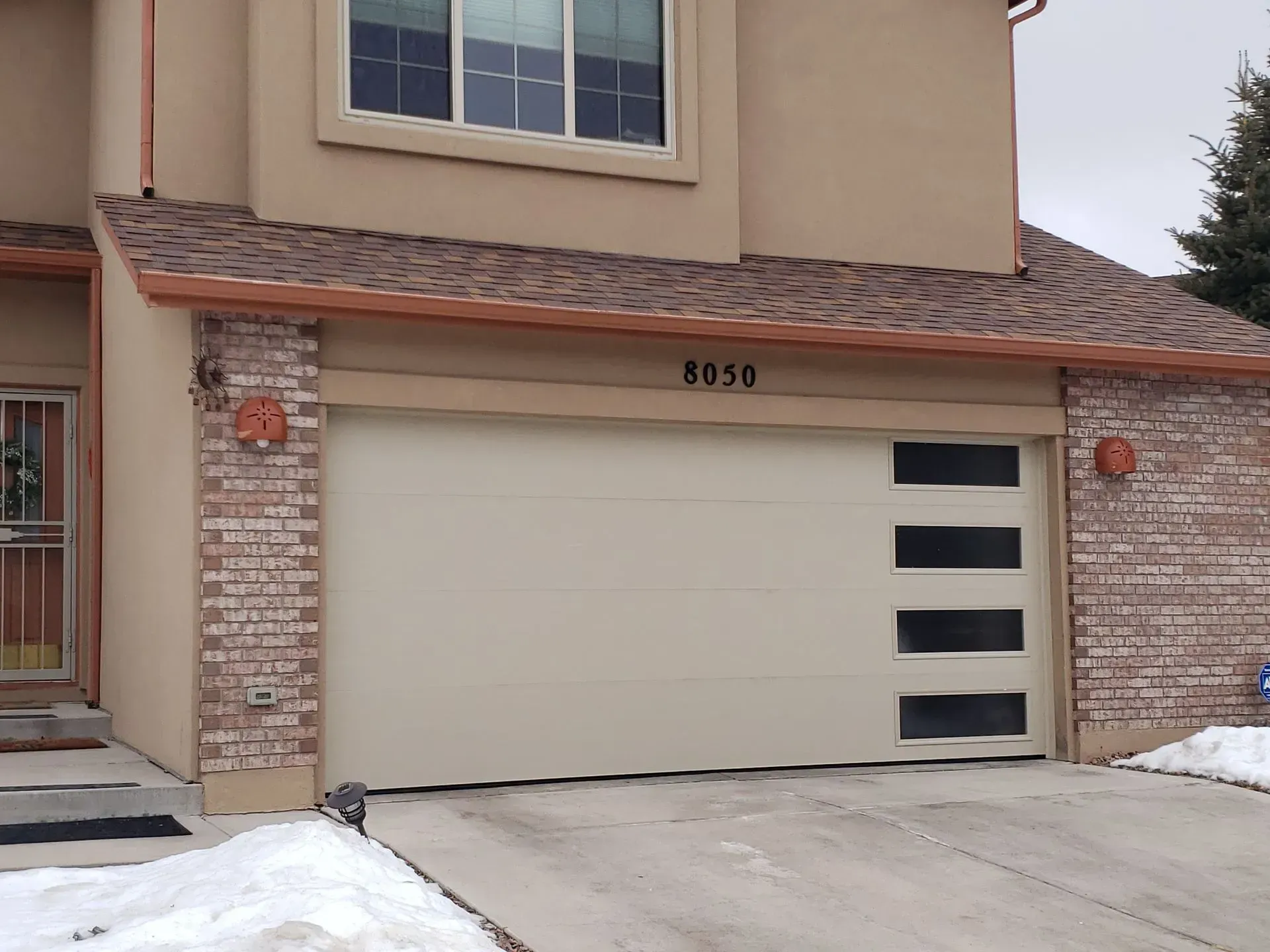 Beige garage door with four rectangular windows; brick facade, tan house, snow.
