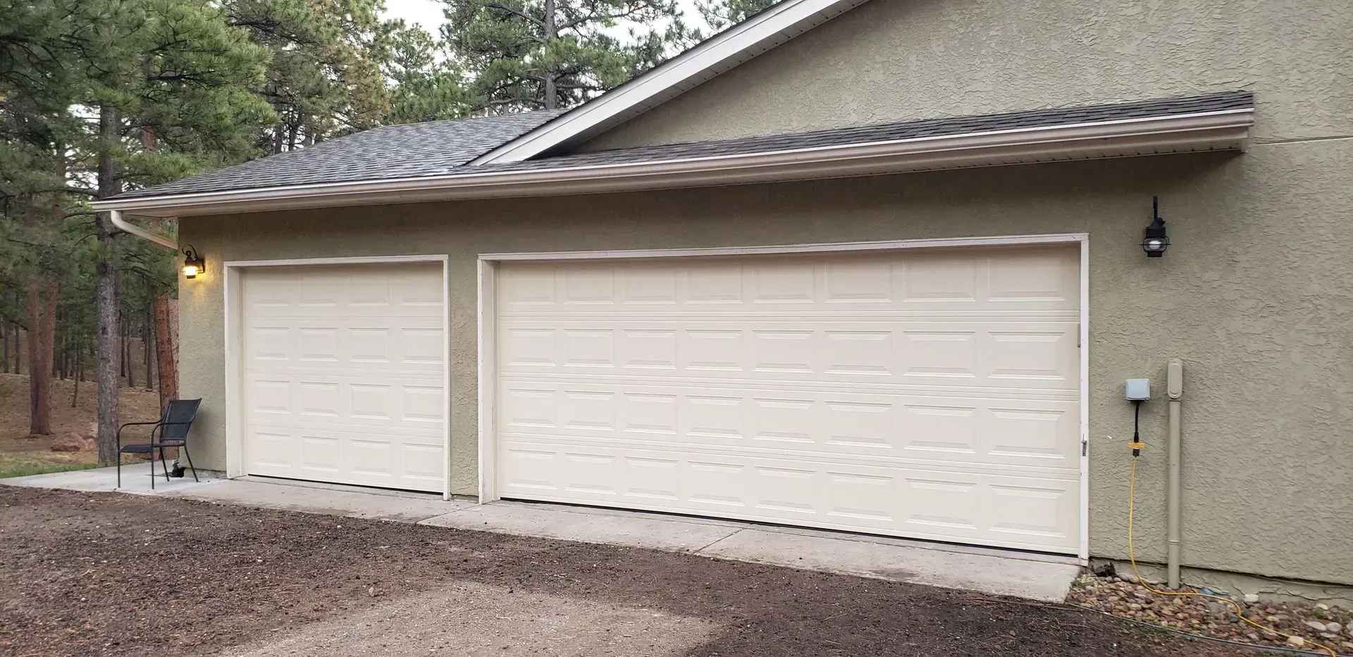 Two-car garage with tan doors and a green exterior, set against a backdrop of trees.