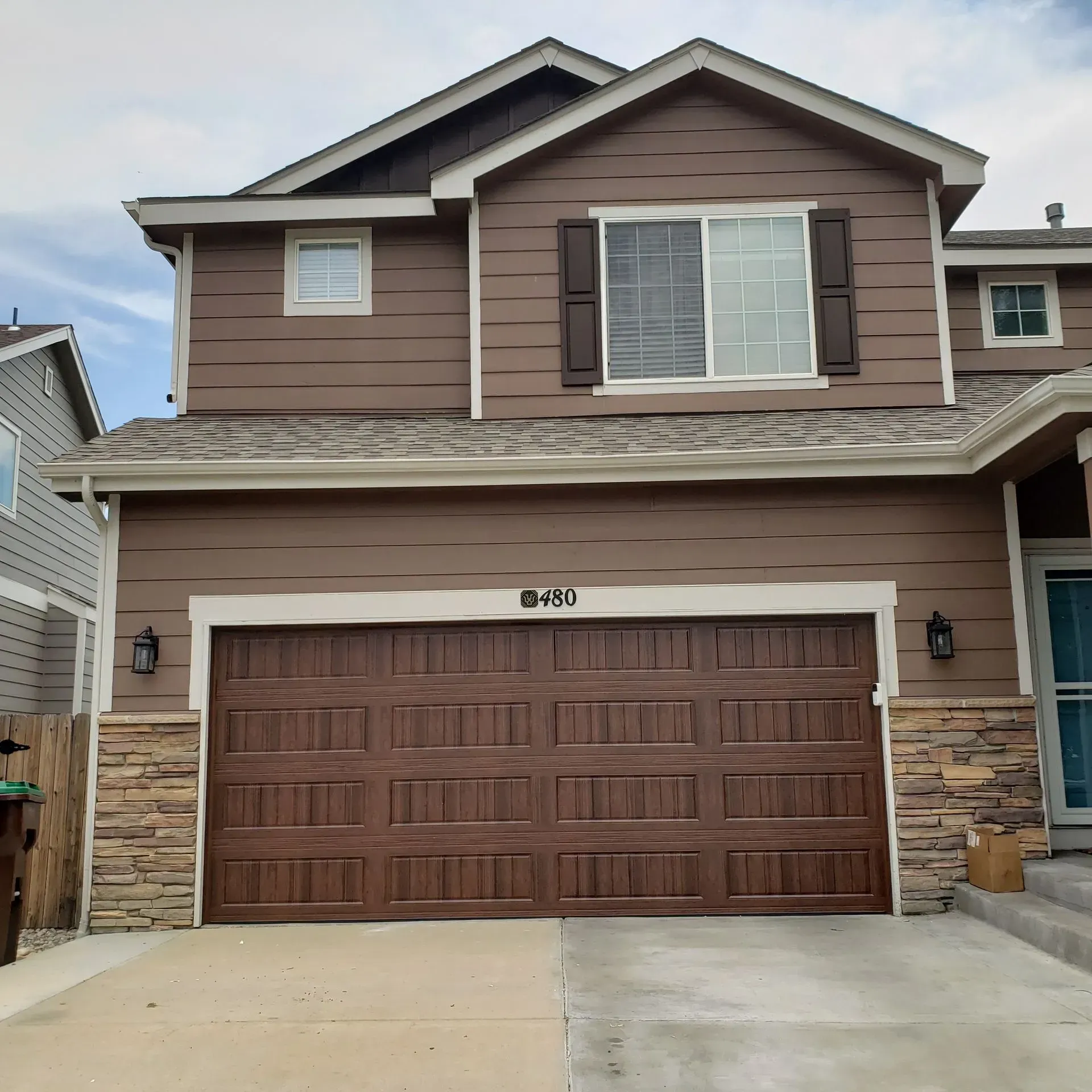 Two-story brown house with a garage door, tan stone accents, and shutters.