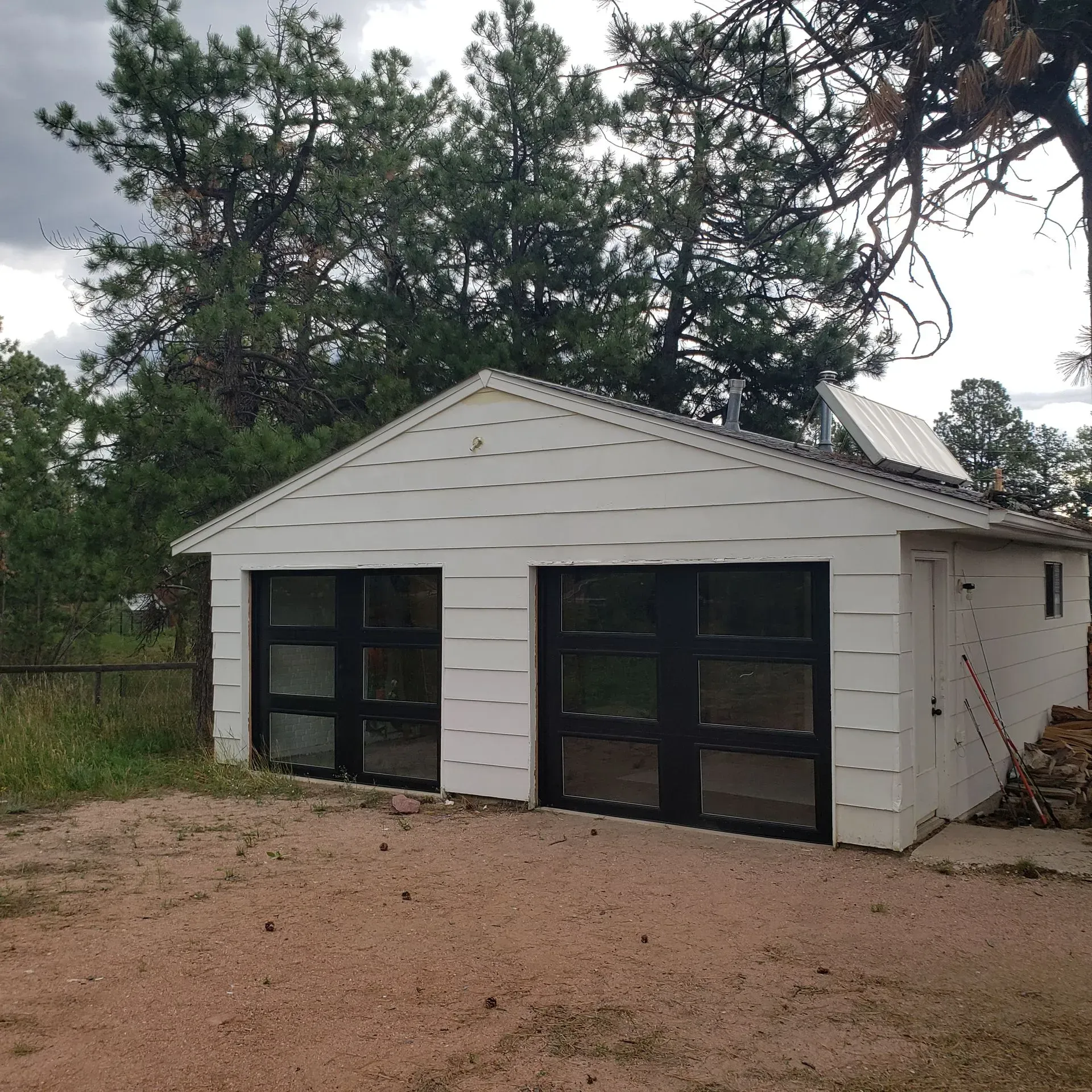 White garage with two black-framed glass doors, set on a dirt driveway, surrounded by trees.
