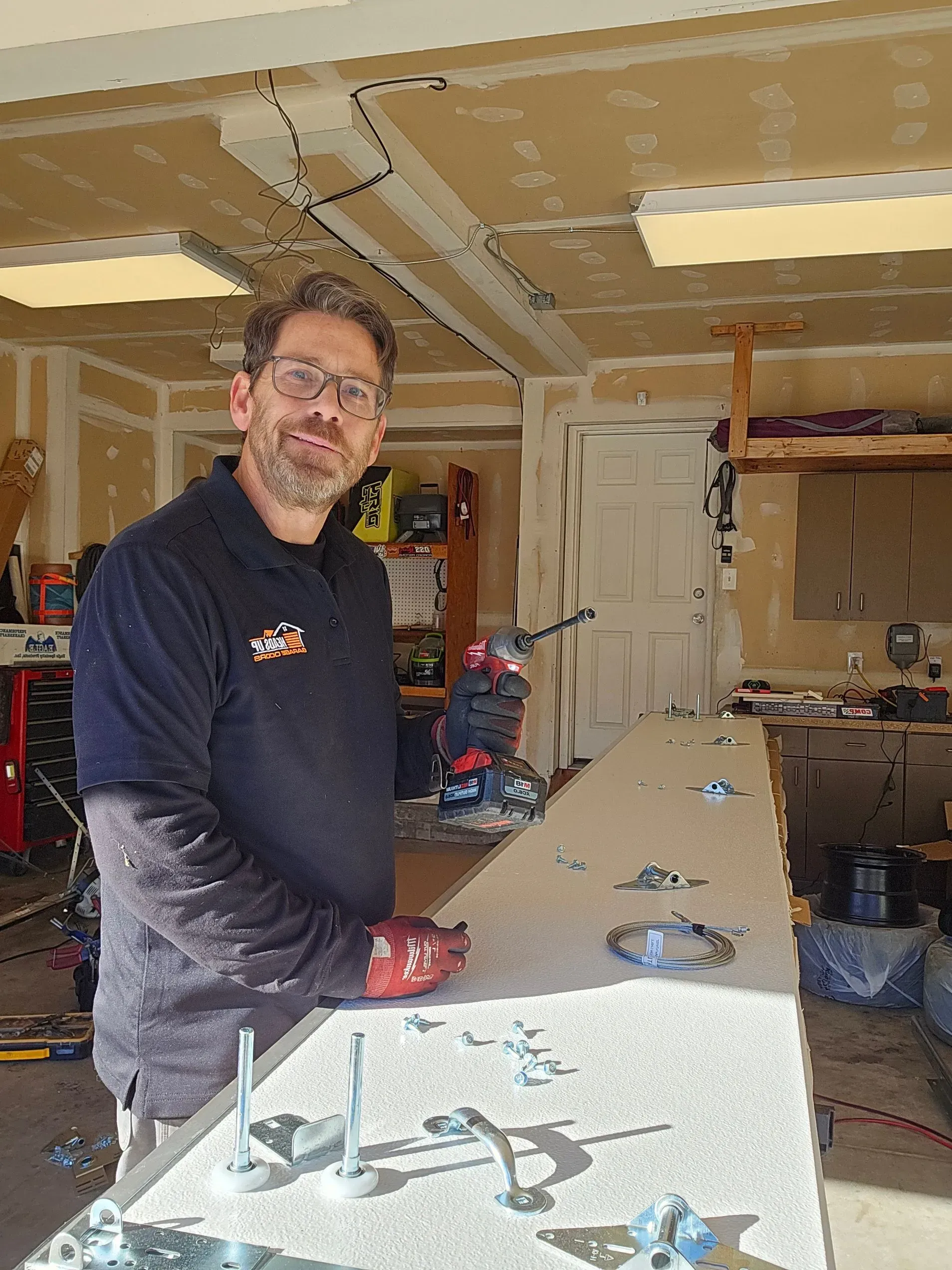 Man working in a garage, smiling, holding a power tool, workbench with metal fixtures.