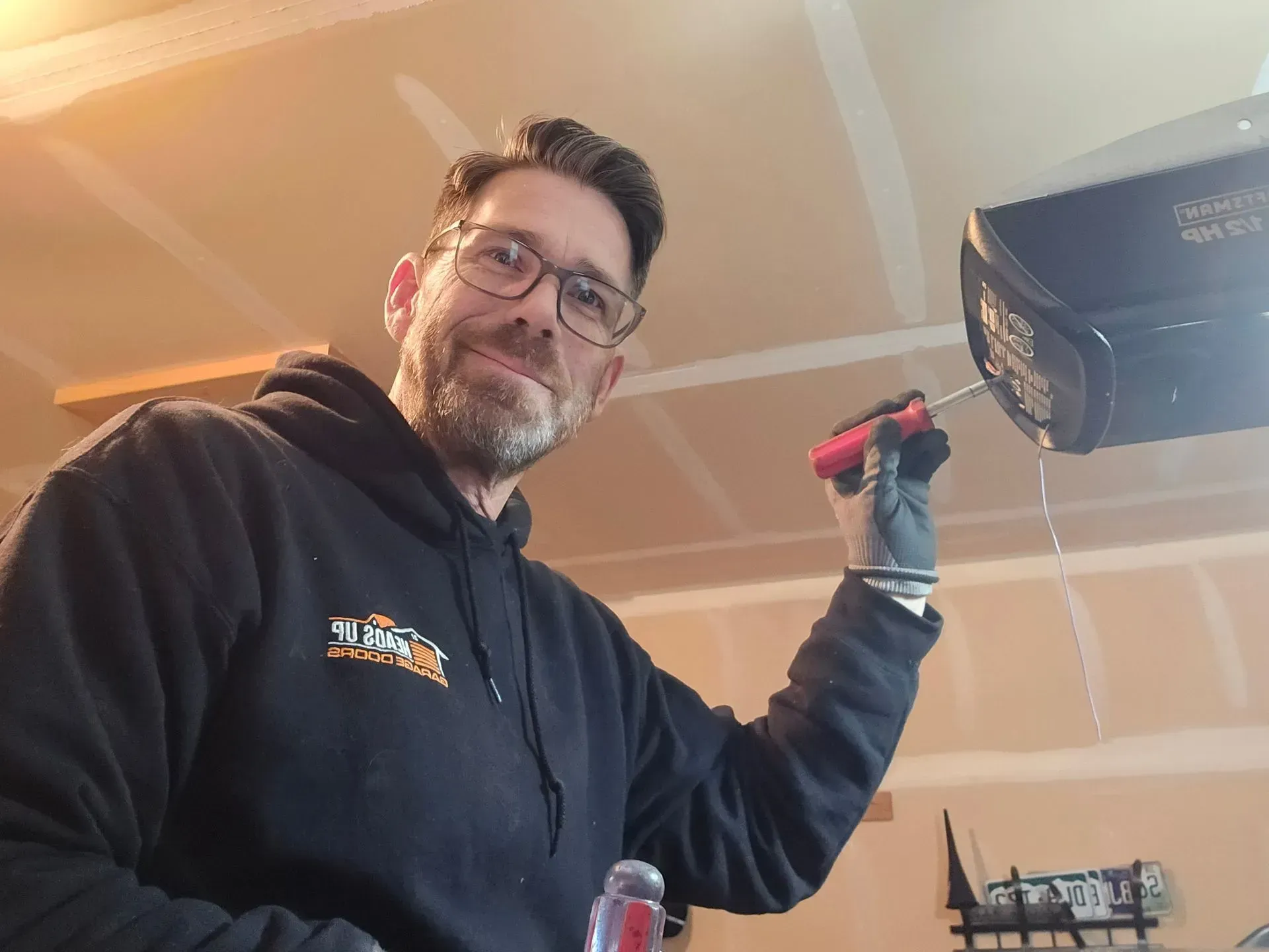Man in a black hoodie fixing a garage door opener with a screwdriver, in a garage.