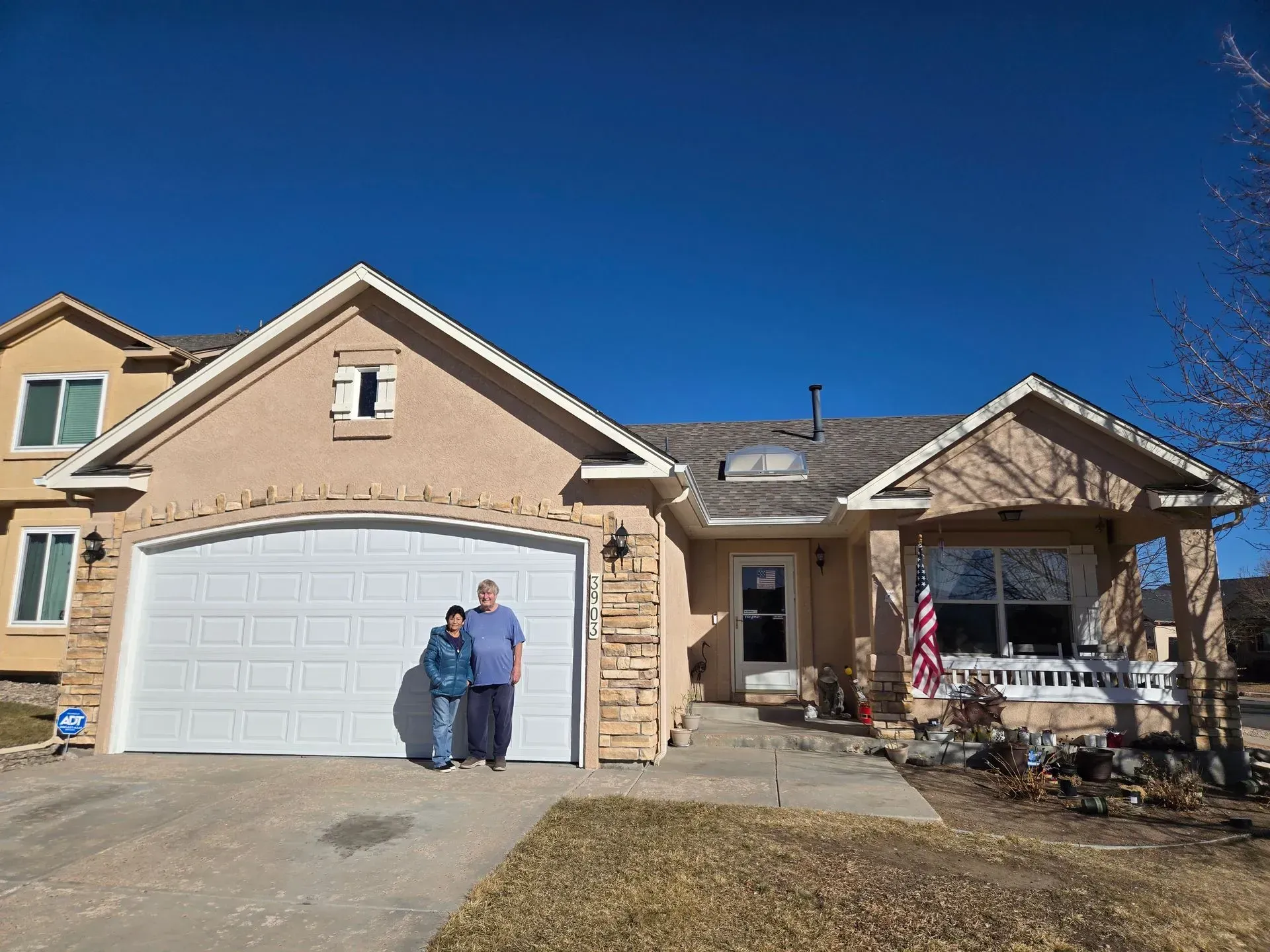 Two people stand in front of a tan house with white garage doors and a blue sky.