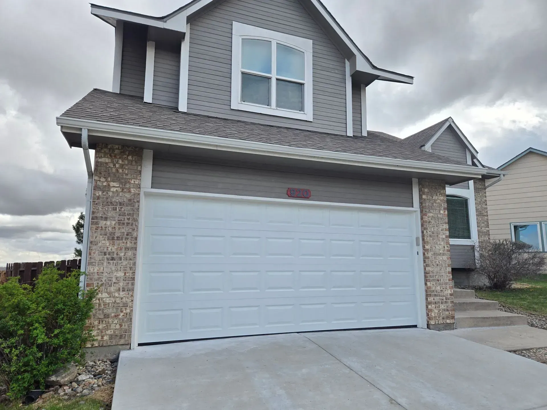 Two-story house with a white garage door, gray siding, and stone accents. Cloudy sky.