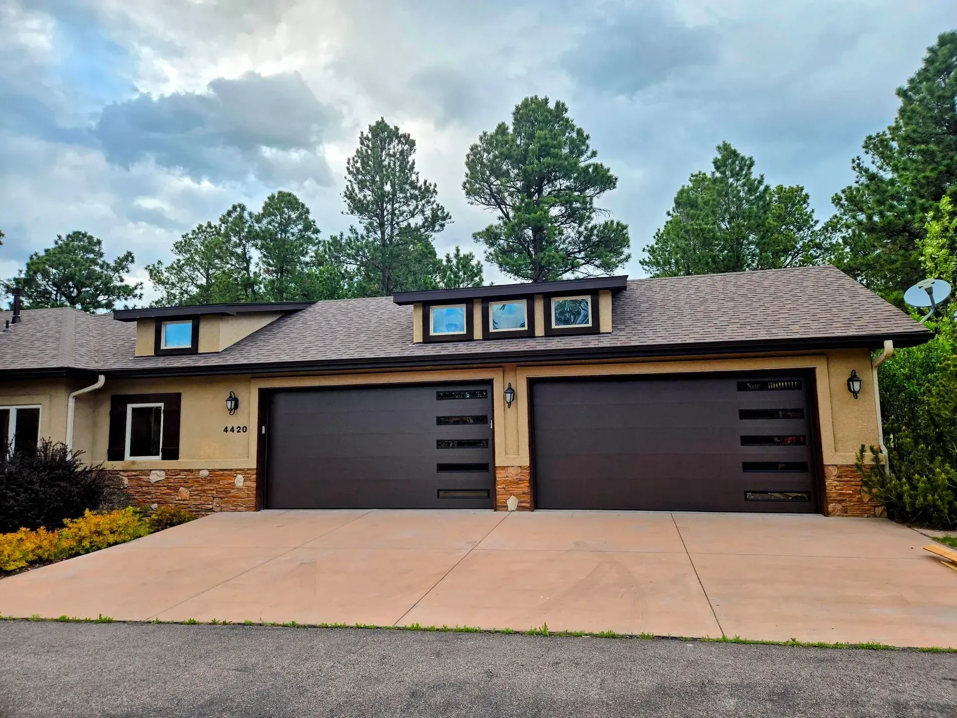 Tan two-car garage with brown doors, stone accents, and dormer windows under a cloudy sky.