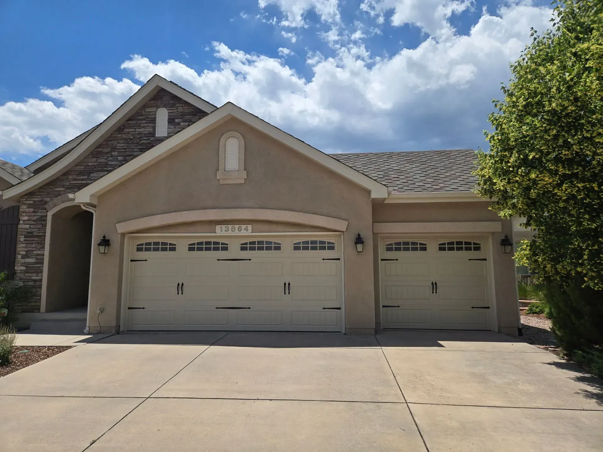 Beige stucco house with a two-car garage, driveway, and a cloudy blue sky.