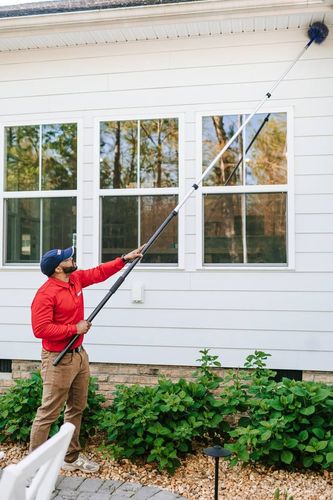 Worker in red uniform spraying pesticide along a house foundation with a backpack sprayer