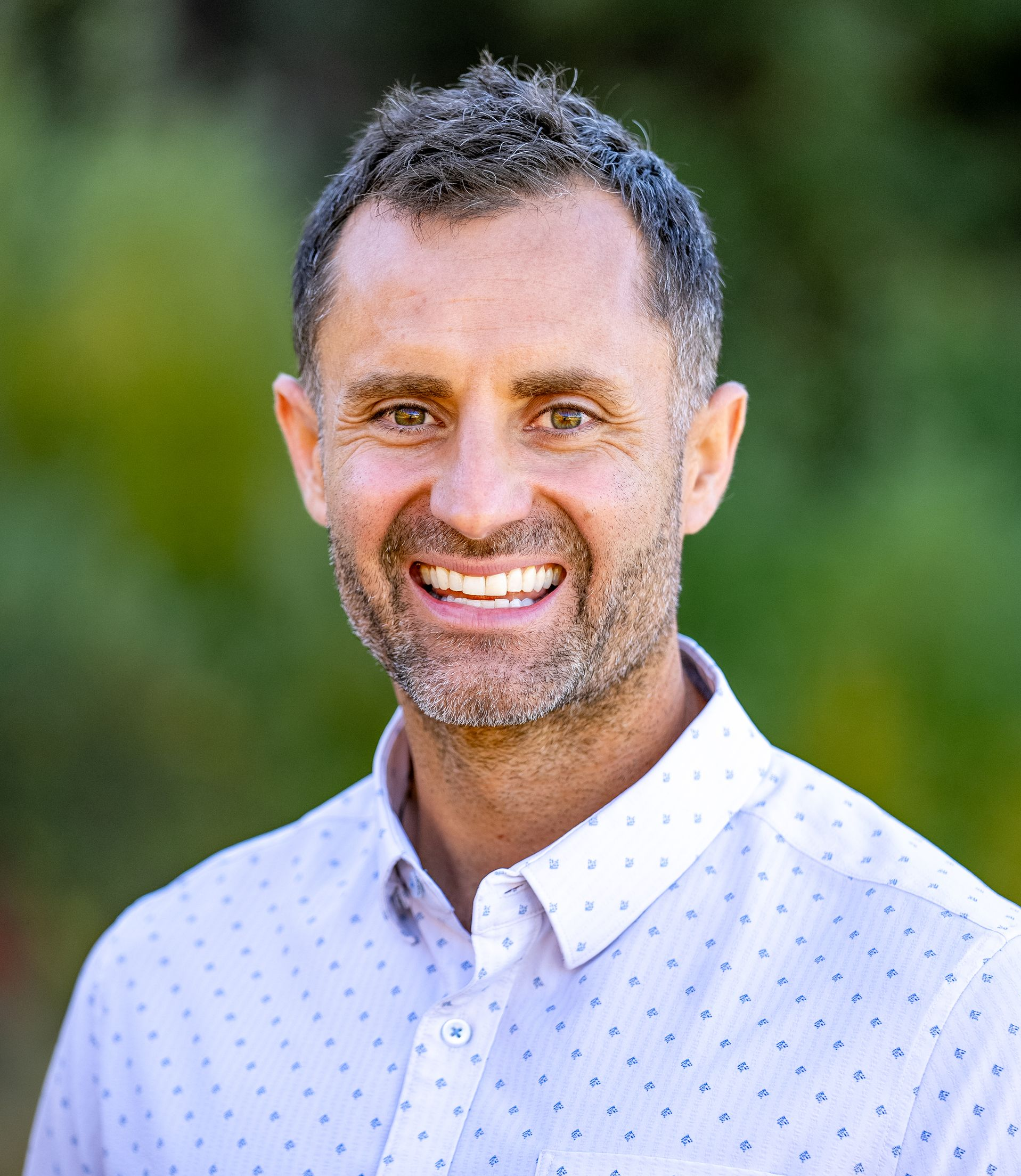 Smiling man in a white patterned shirt outdoors with a blurred green background