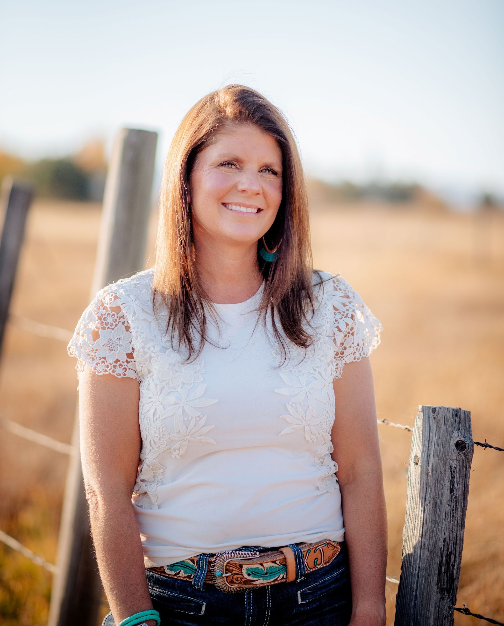 Woman standing by a fence in a sunlit field, wearing a white lace top and smiling at the camera.