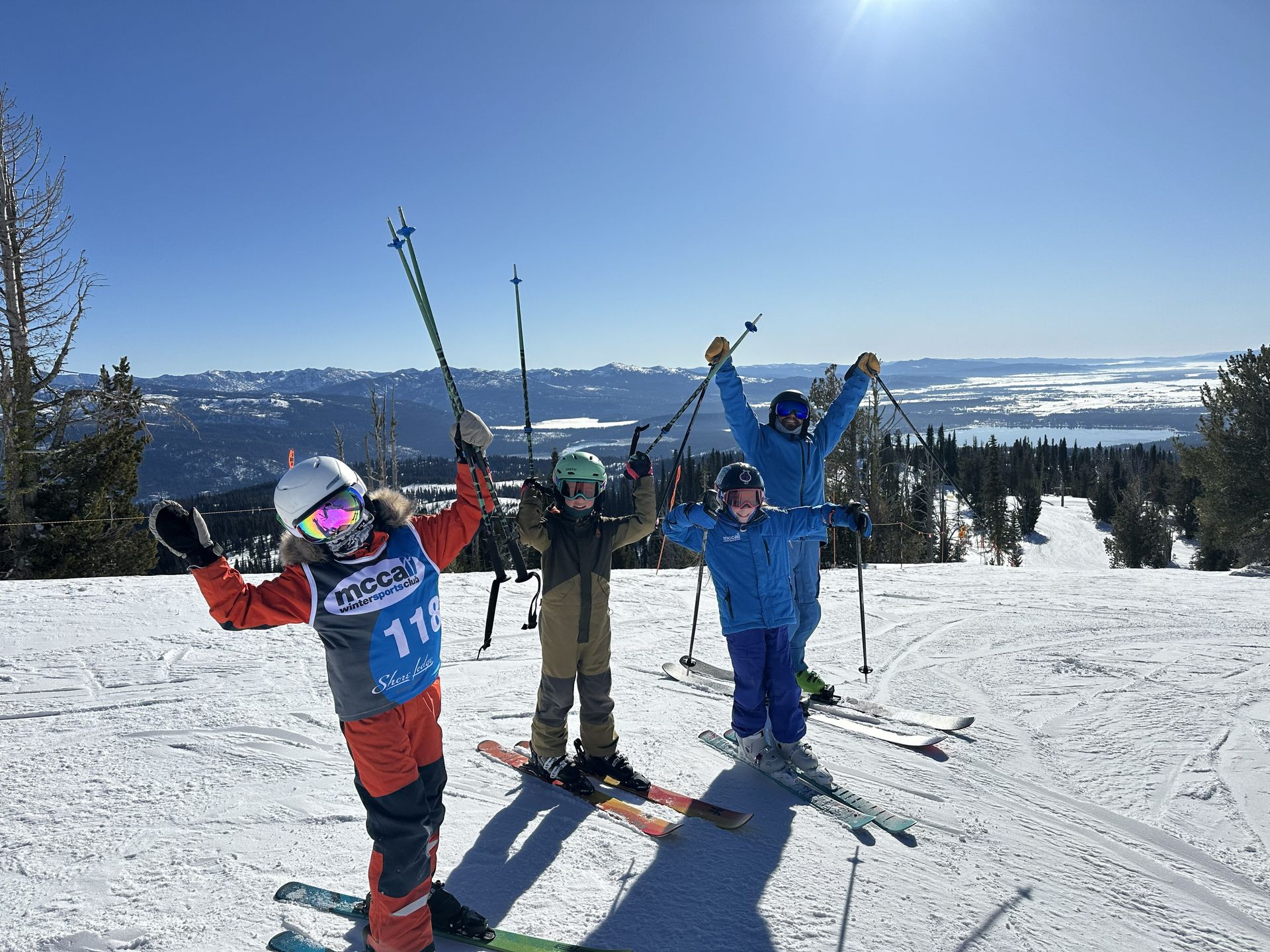 Three skiers in colorful gear pose with skis raised on a snowy mountain under a clear blue sky.