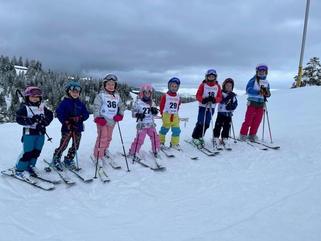 Children posing with skis on a snowy slope, wearing colorful winter gear and helmets.