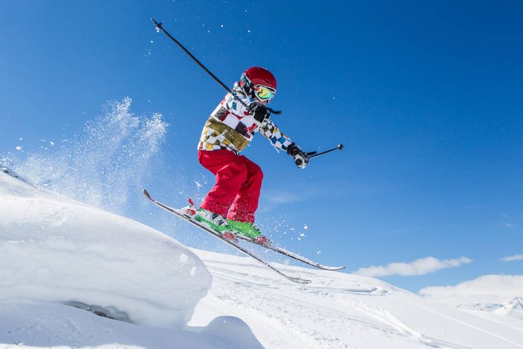 Skier in red pants and helmet jumps through powder on a bright snowy mountain slope.