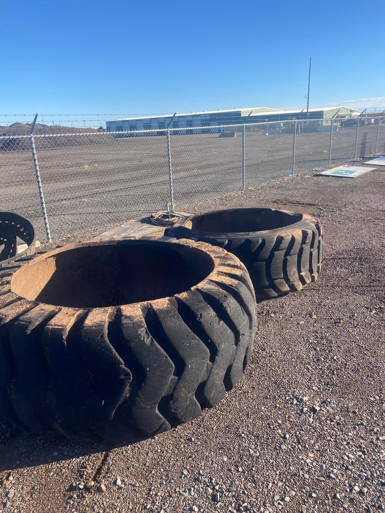 Two large, worn tires with open centers sit on gravel. A chain-link fence and buildings are visible in the background.