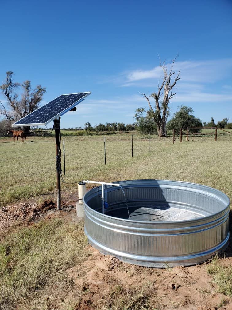 Solar panel powering a water trough in a field. Brown and green landscape with a blue sky.