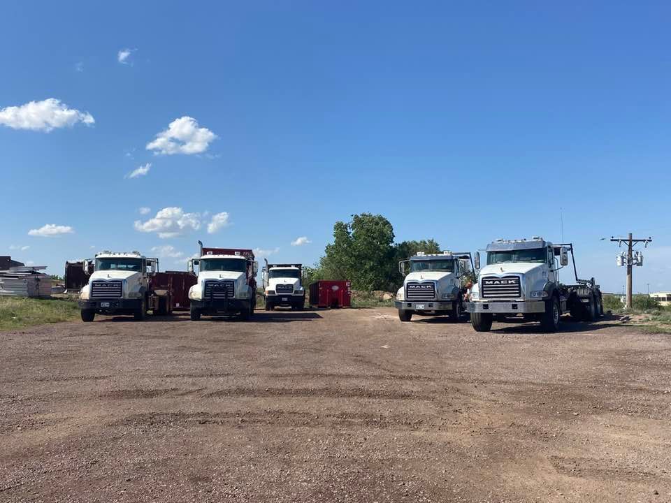 Five white semi-trucks parked on a gravel lot under a blue sky with some clouds.