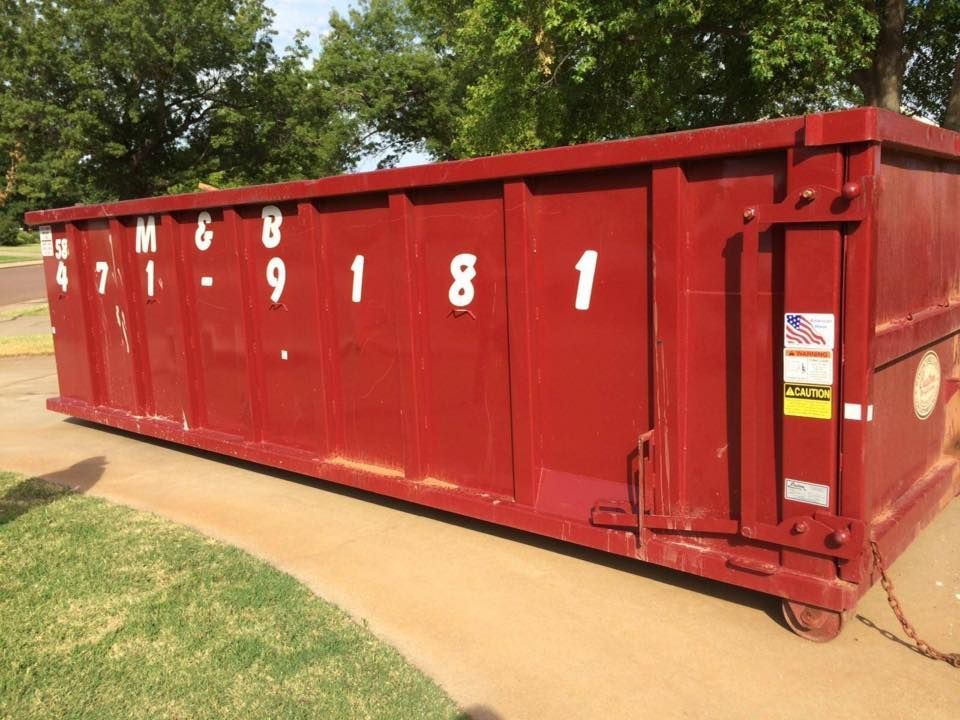 Red dumpster on a concrete surface with the numbers 471-9181. Green grass and trees in background.
