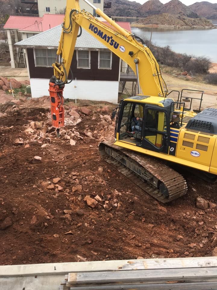 Yellow Komatsu excavator using a jackhammer attachment on reddish soil. A house and water are in the background.