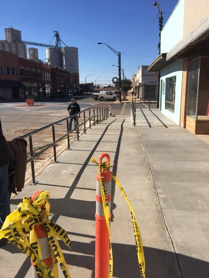 Sidewalk with ramp under construction, orange cones, yellow caution tape. Buildings and grain silos in the distance.