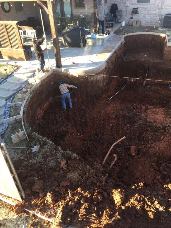 Workers excavating a curved pool; one person in the hole, others above. Soil, concrete, and building in the background.