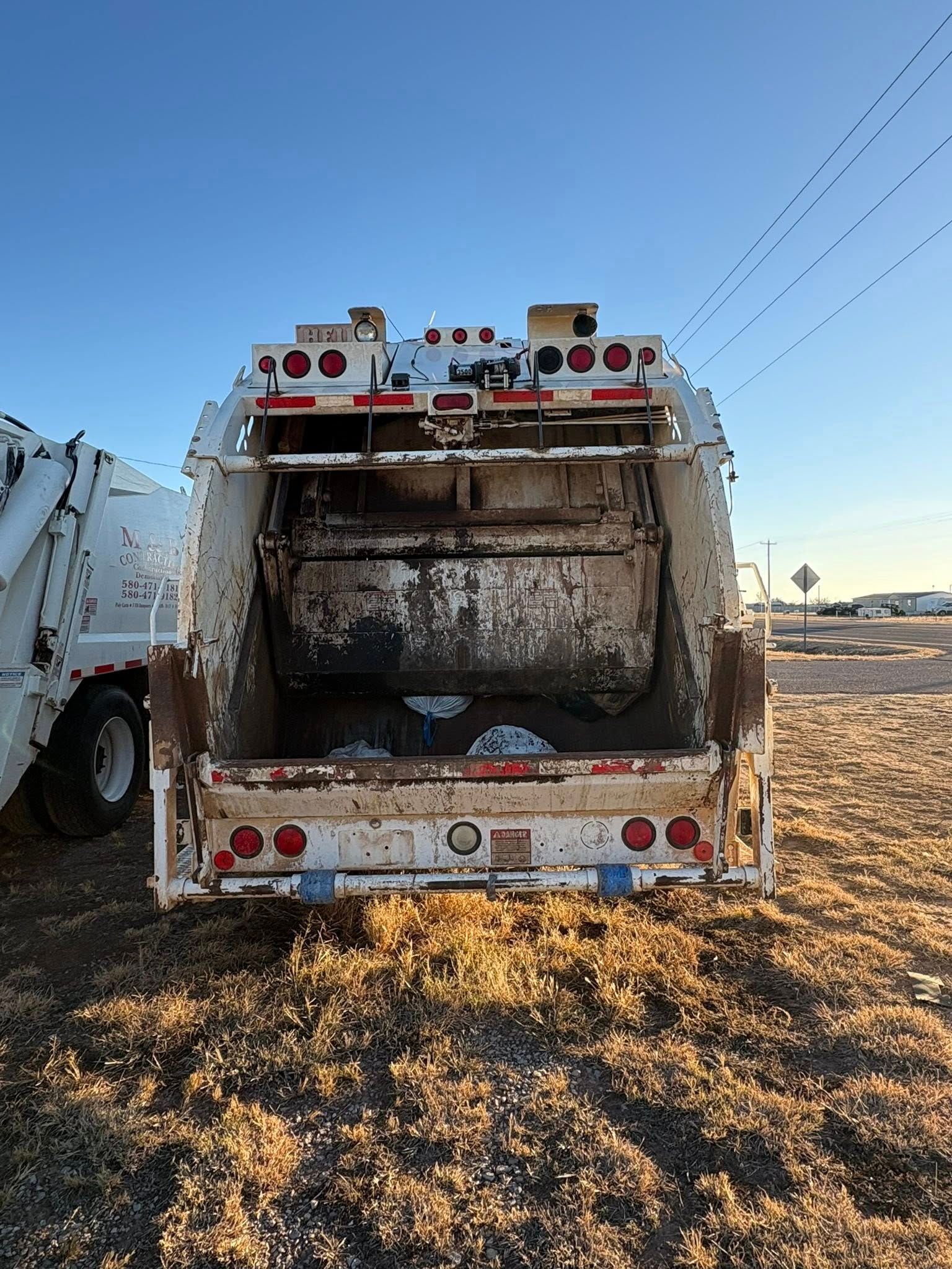 Rear view of a white, dirty garbage truck with its container open on a field, blue sky in background.