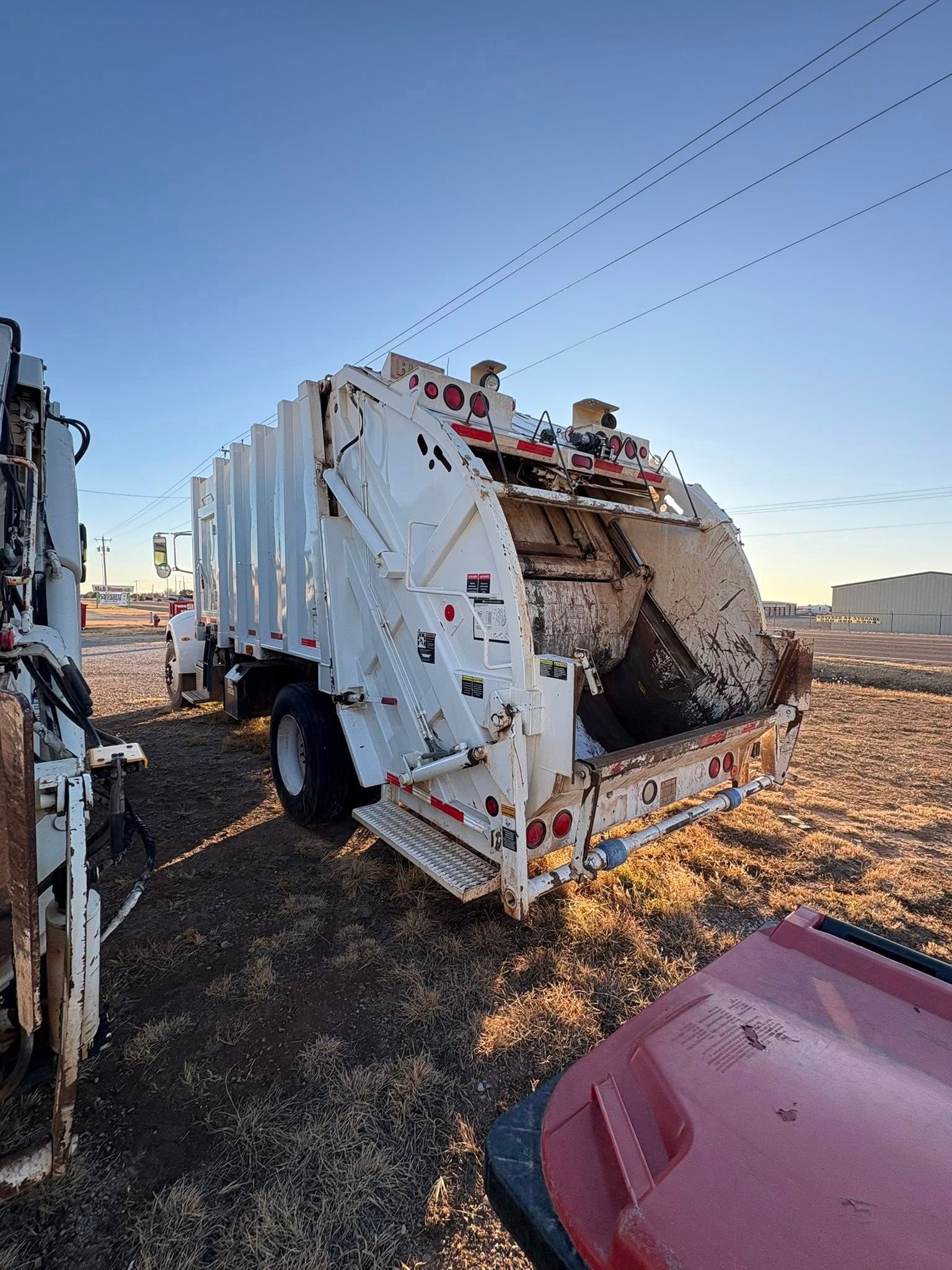 White garbage truck on brown ground, wires overhead, red car partially in view.